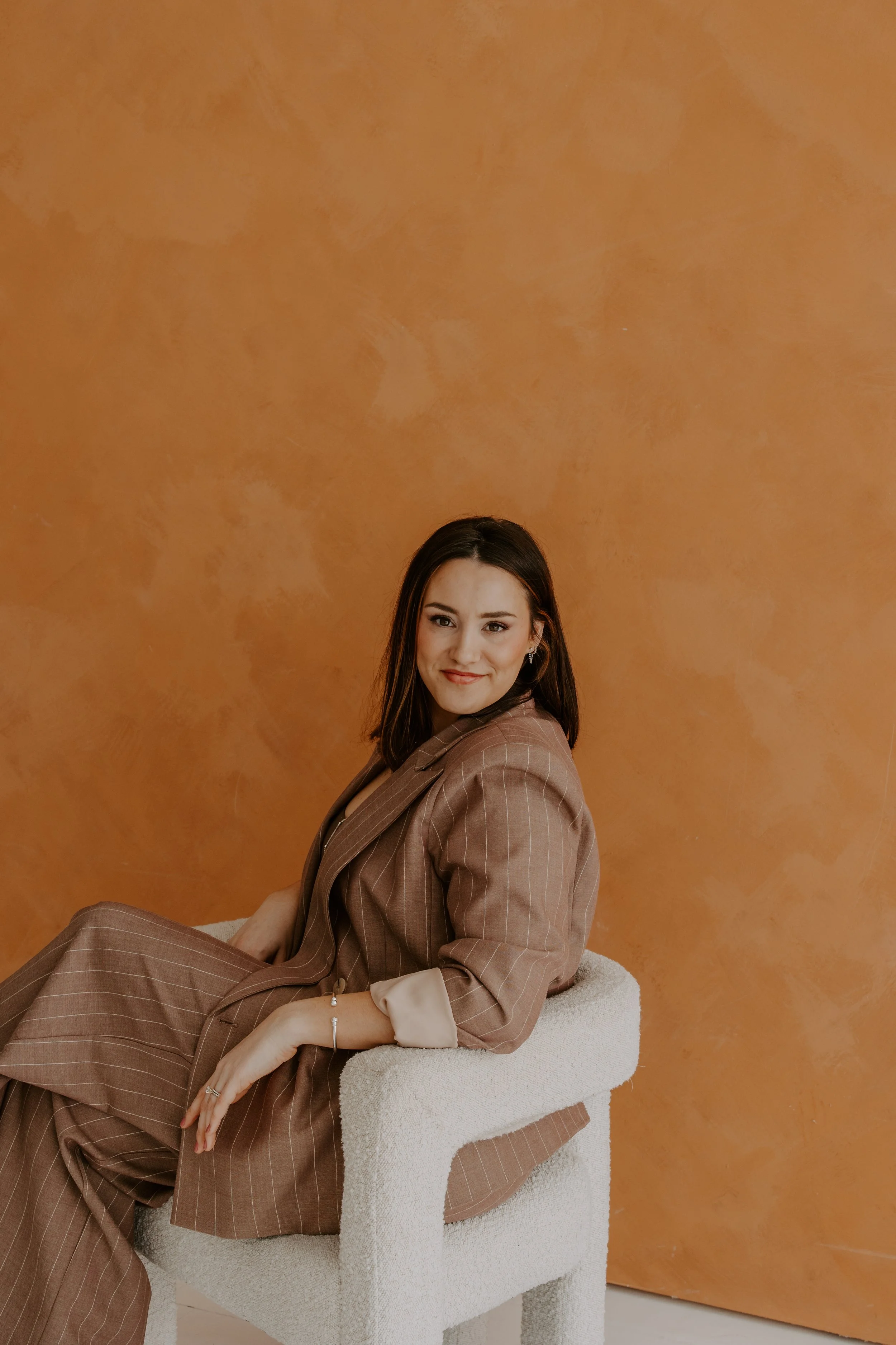 A woman with dark brown hair, smiling, wearing a brown pinstripe suit, sitting on a white textured armchair against an orange textured wall.