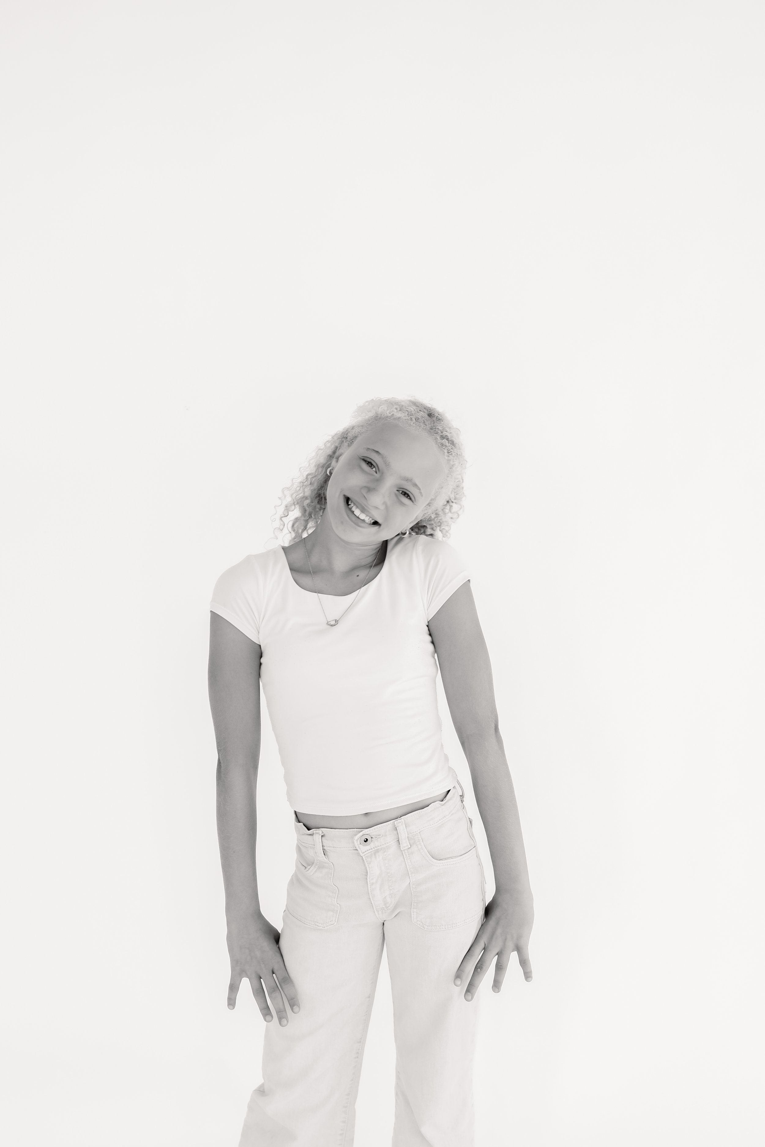 A young girl with curly hair smiling and tilting her head to the side, standing against a plain white background.