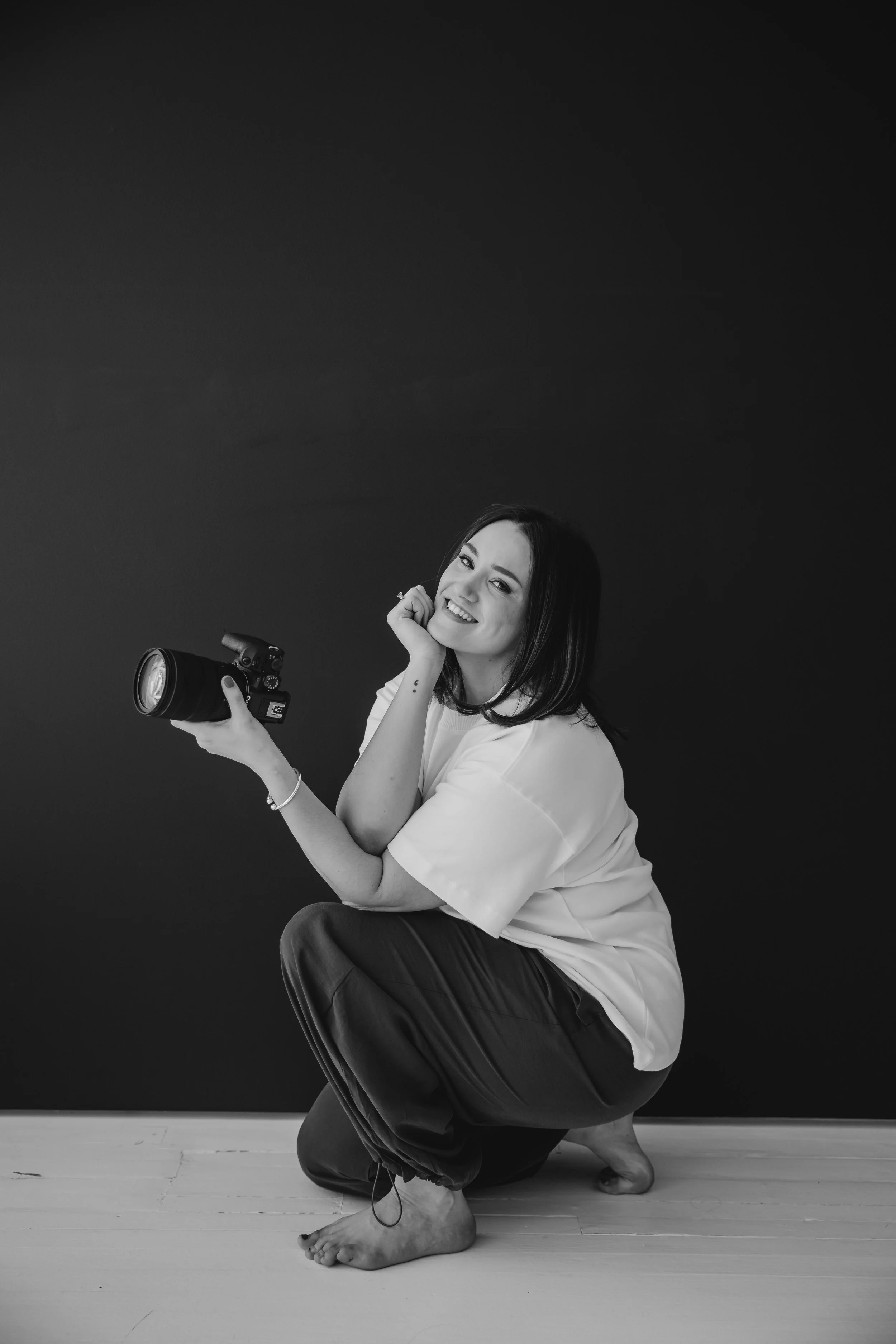 A woman with dark hair, sitting on the floor, holding a camera and smiling at the camera, with a plain dark background.