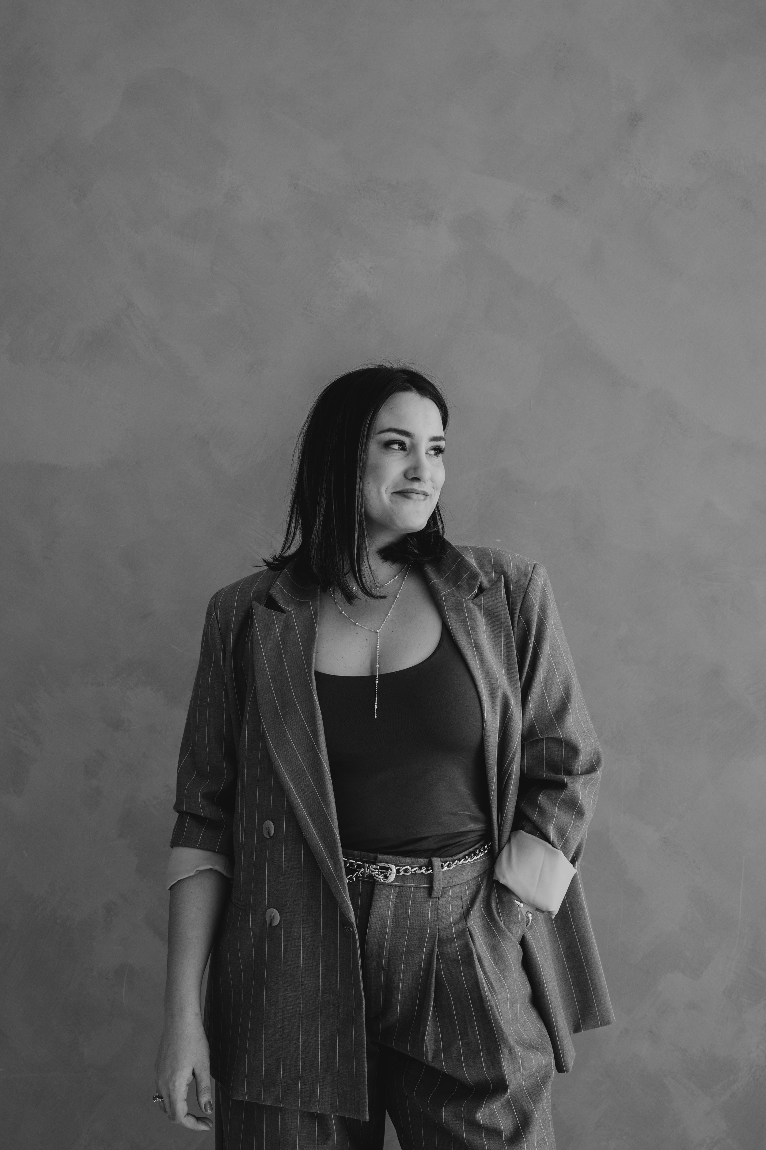Black and white photo of a woman in a pinstripe suit with a relaxed pose, standing against a plain textured wall, smiling slightly and looking to her left.