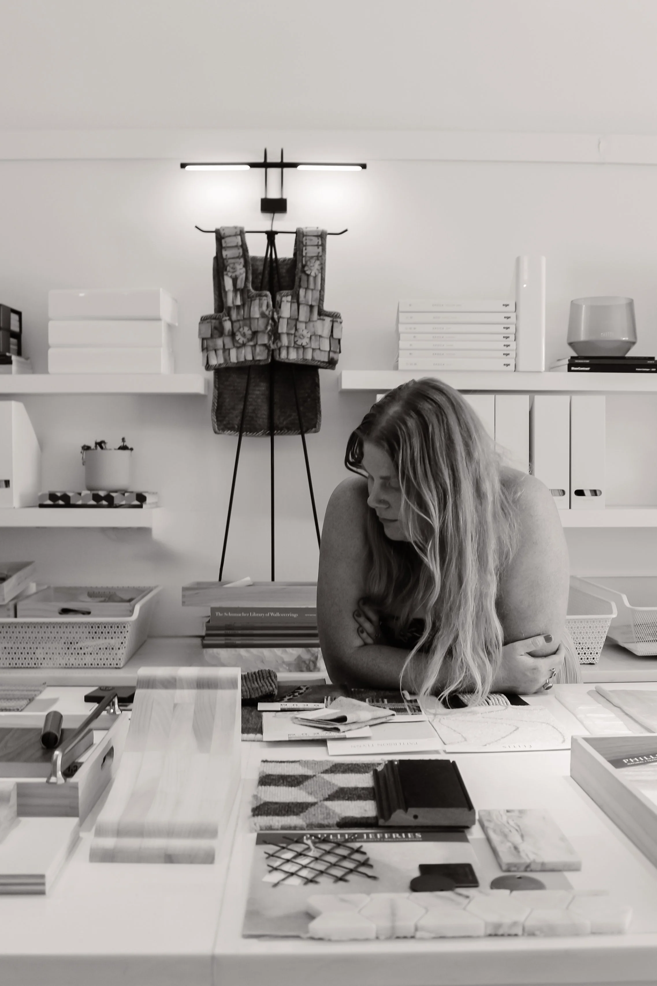 A woman with long hair is leaning forward on a table, studying various design and interior decorating materials and samples in a well-organized room with white shelves and decorative objects.