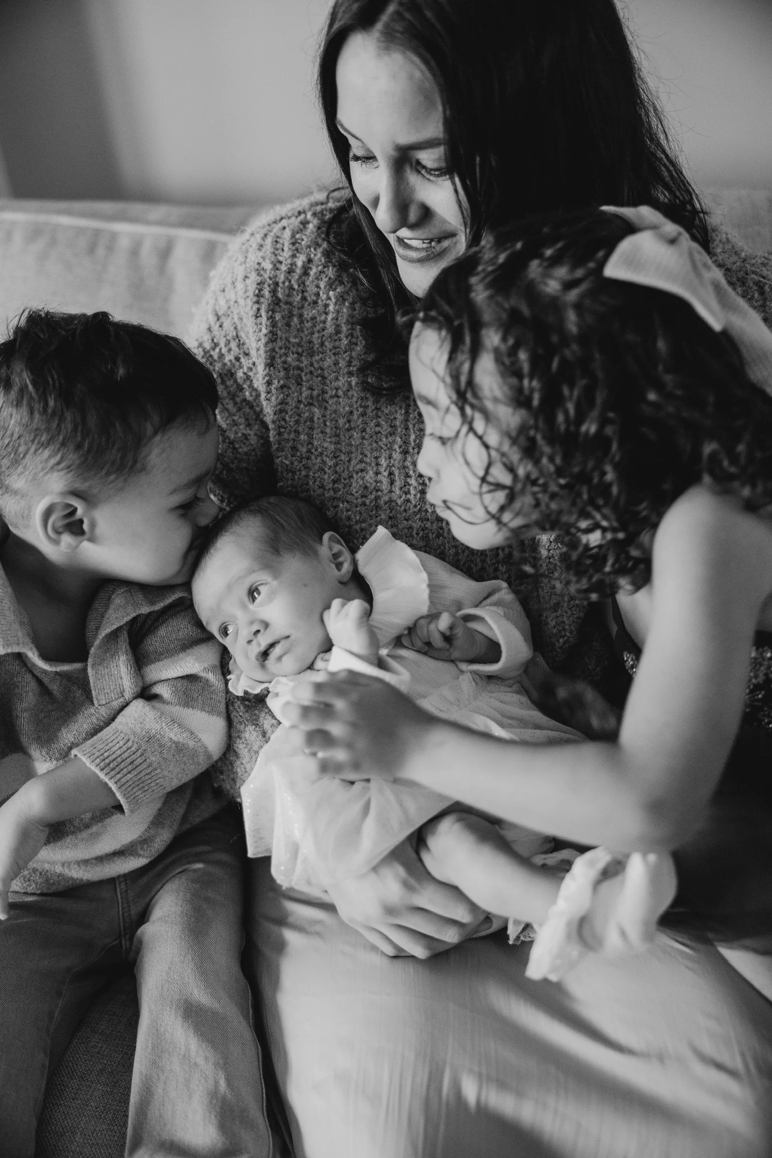 A woman with two children holding a baby and comforting her, with another young child leaning in, all on a sofa.
