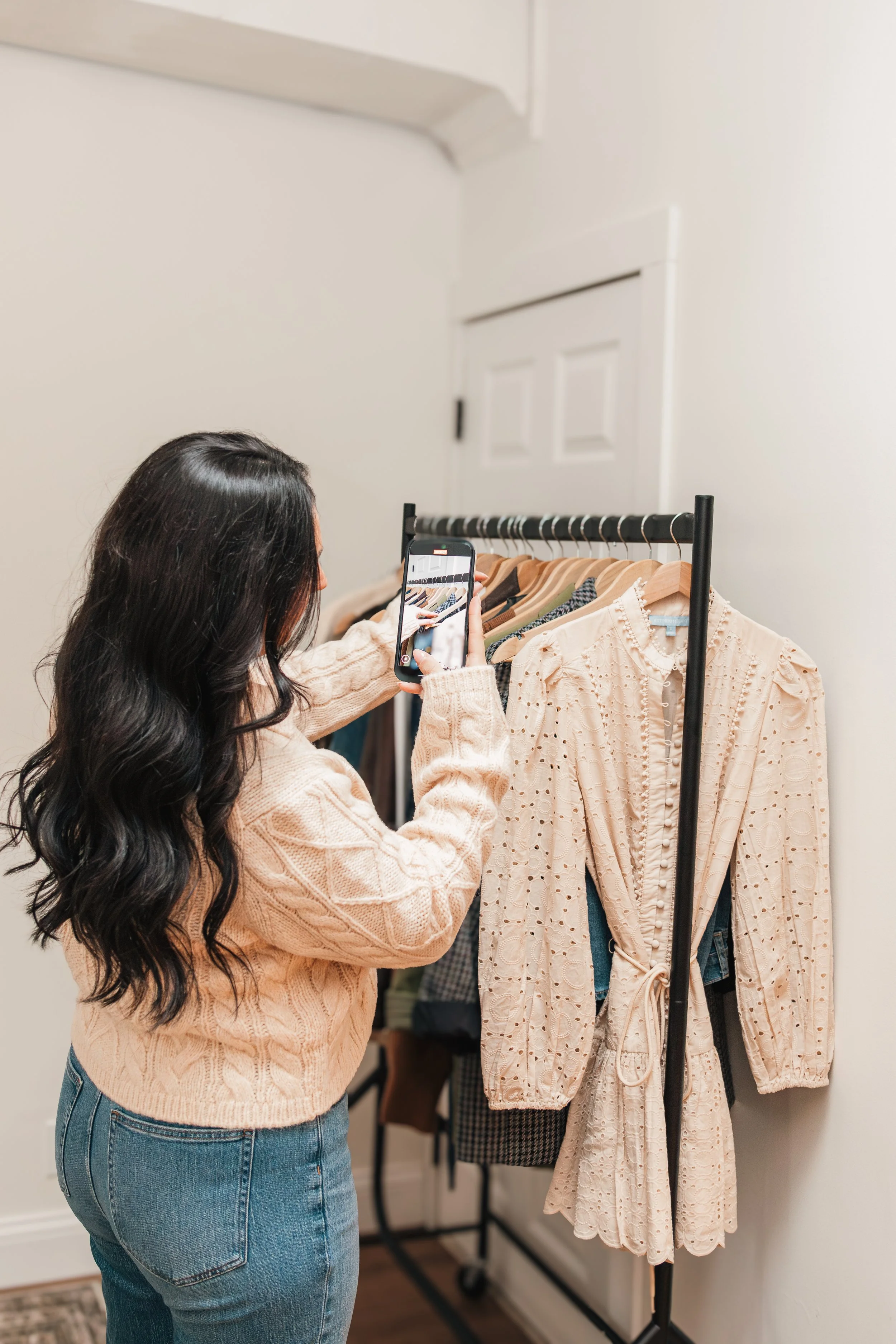 A woman with long dark hair wearing a beige sweater taking a photo of a clothing rack with her smartphone.