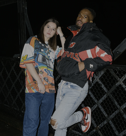Two young adults, a woman and a man, standing on a bridge at night with a dark sky in the background. The woman has long brown hair and is wearing a colorful patterned shirt and blue jeans. The man has short hair and a beard, wearing a black and red jacket, light distressed jeans, and red and white Nike sneakers.