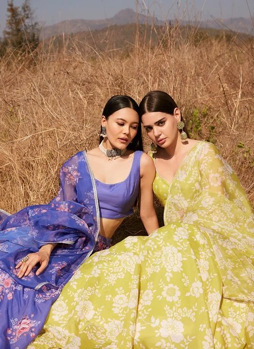 Two women sitting in a dry, grassy field with mountains in the background, wearing colorful traditional Indian dresses and jewelry.