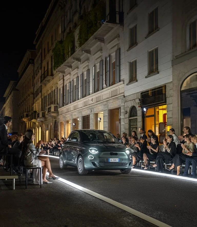 A compact black car drives down a runway on a city street at night, with a crowd dancing and watching on both sides, illuminated by streetlights and nearby buildings.