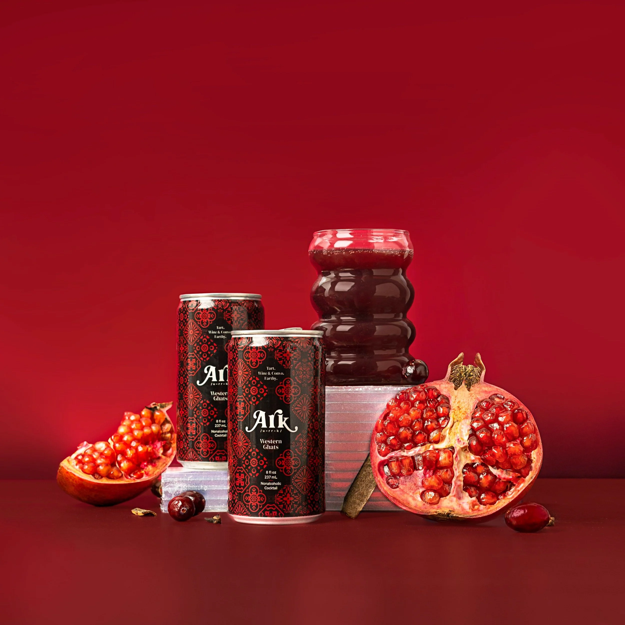 Pomegranate and two cans of Arki Western Grape juice with a container of grape jelly and a cinnamon stick on a red background.