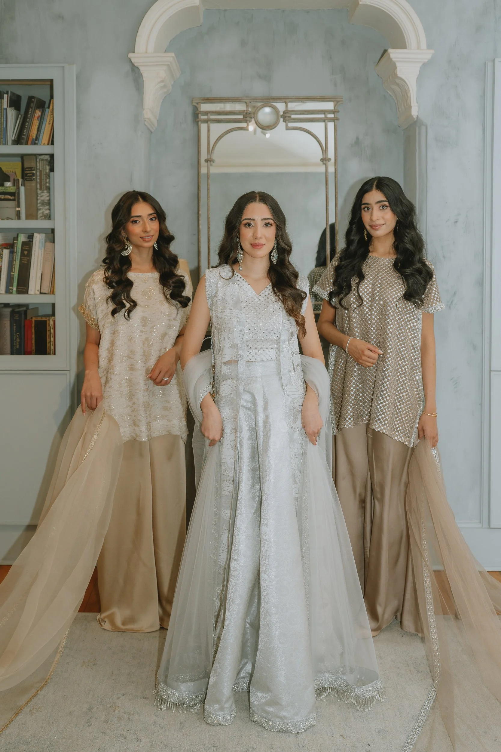 Three women in elegant attire standing in front of a mirror with a light-colored wall background.