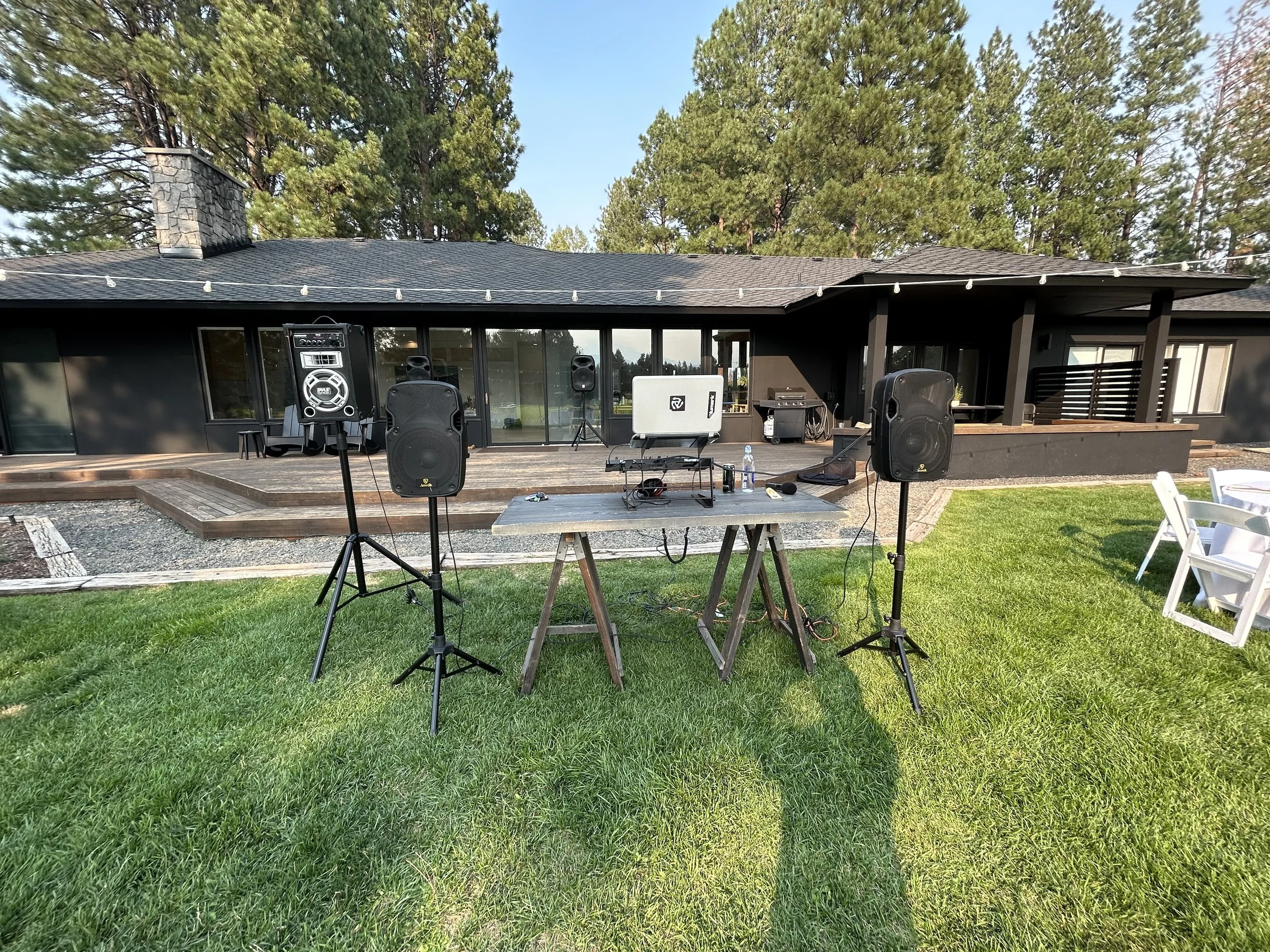 Backyard with a DJ setup on the lawn, including speakers, a DJ controller, and a laptop, in front of a modern house with large glass doors and a wooden deck, surrounded by trees.