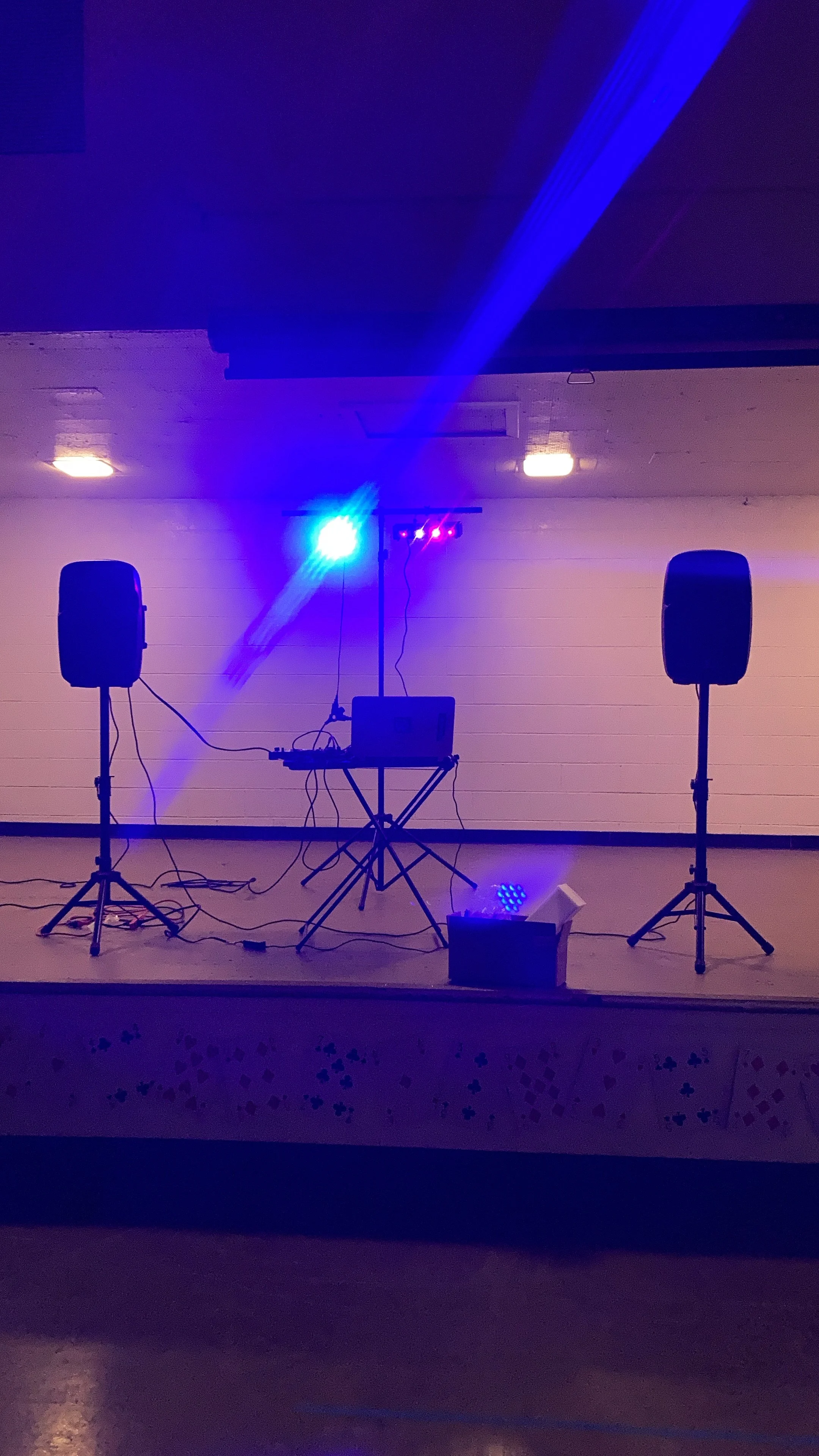 A stage with two black speakers on stands, colorful lighting, and a DJ setup with a laptop and lighting equipment, set against a plain wall.