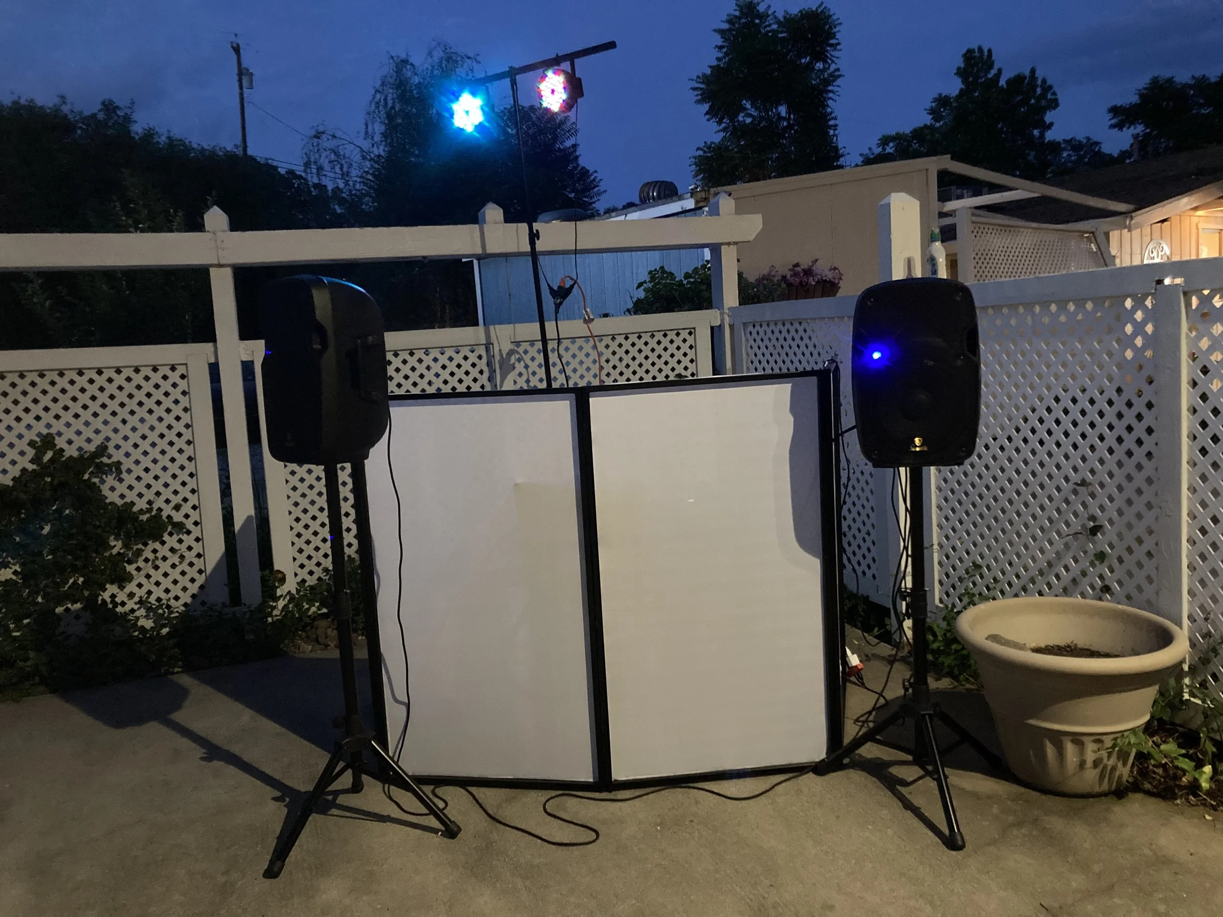 An outdoor DJ setup featuring two black speakers on stands, a white folding screen, and colorful lights on a tall stand, set against a white fence and a yard at dusk.