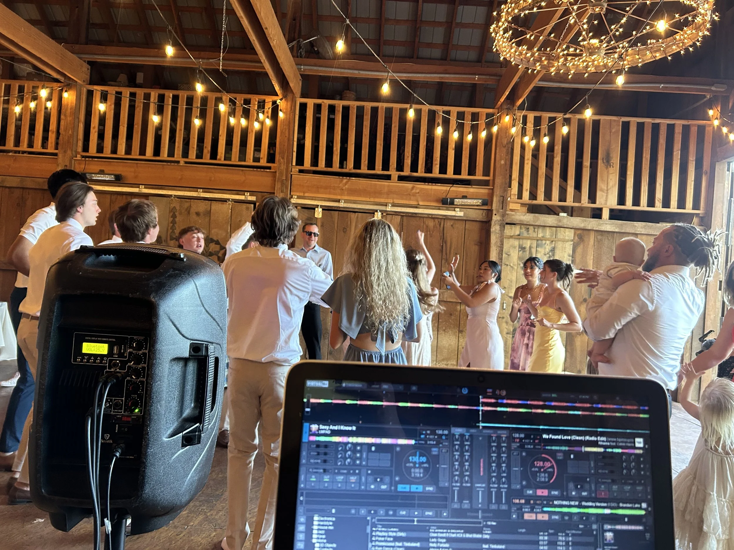 People dancing and socializing at a wedding reception in a wooden barn with string lights and a chandelier, with DJ equipment and a laptop in the foreground.