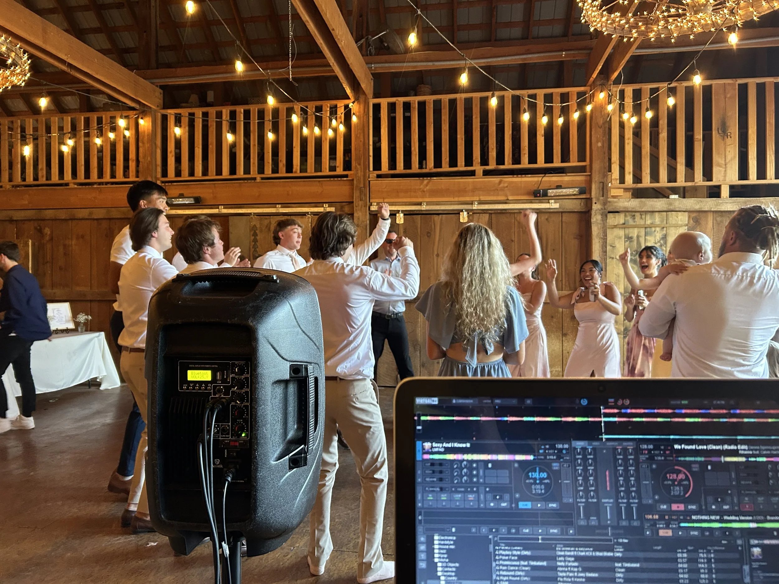 People dancing at a celebration event in a rustic wooden venue with string lights, with DJ equipment and a laptop in the foreground.