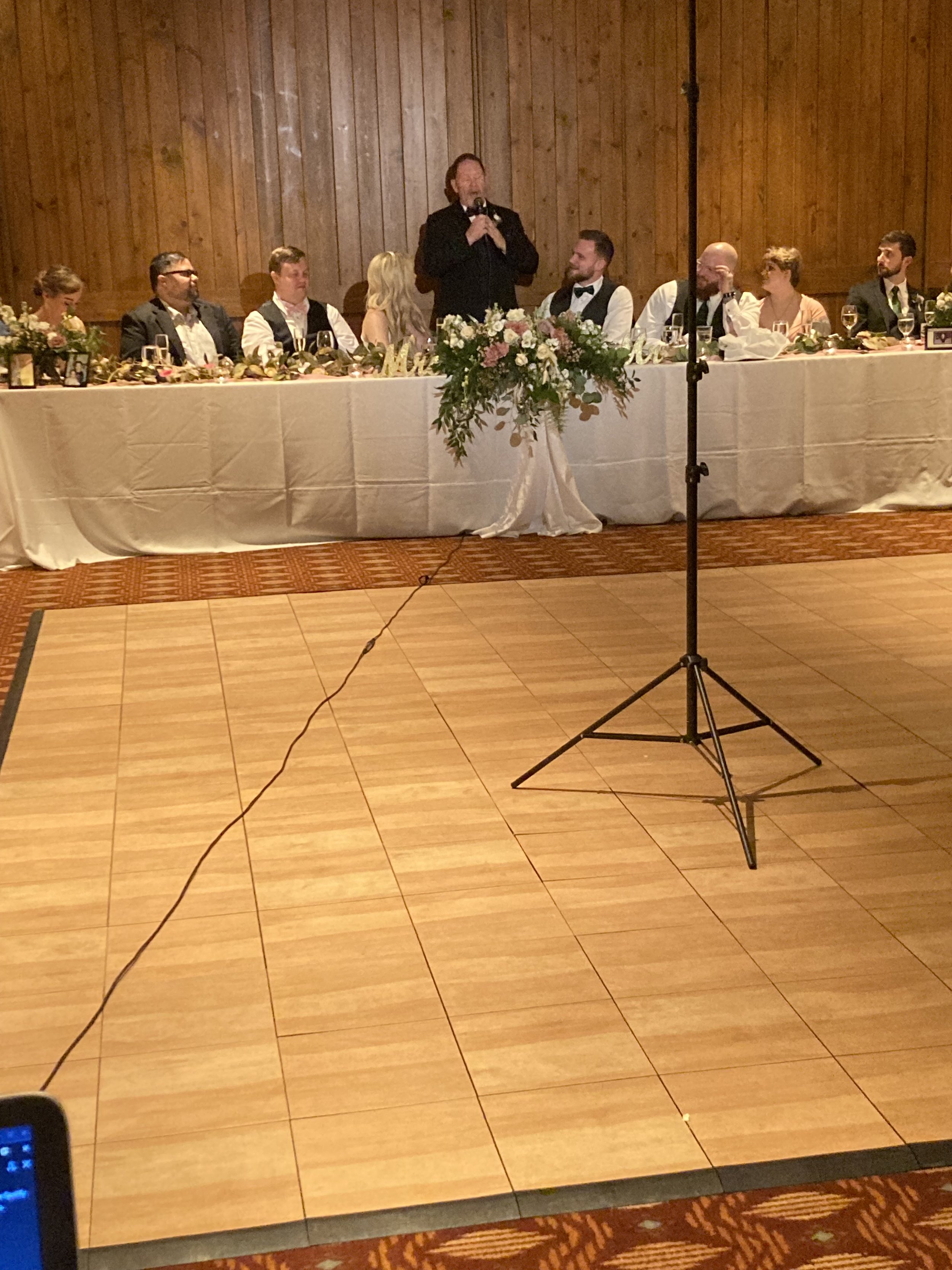 A woman giving a speech at a wedding reception table decorated with flowers and a white tablecloth, with guests seated beside her. A microphone stand is in front of the table, on a dance floor.