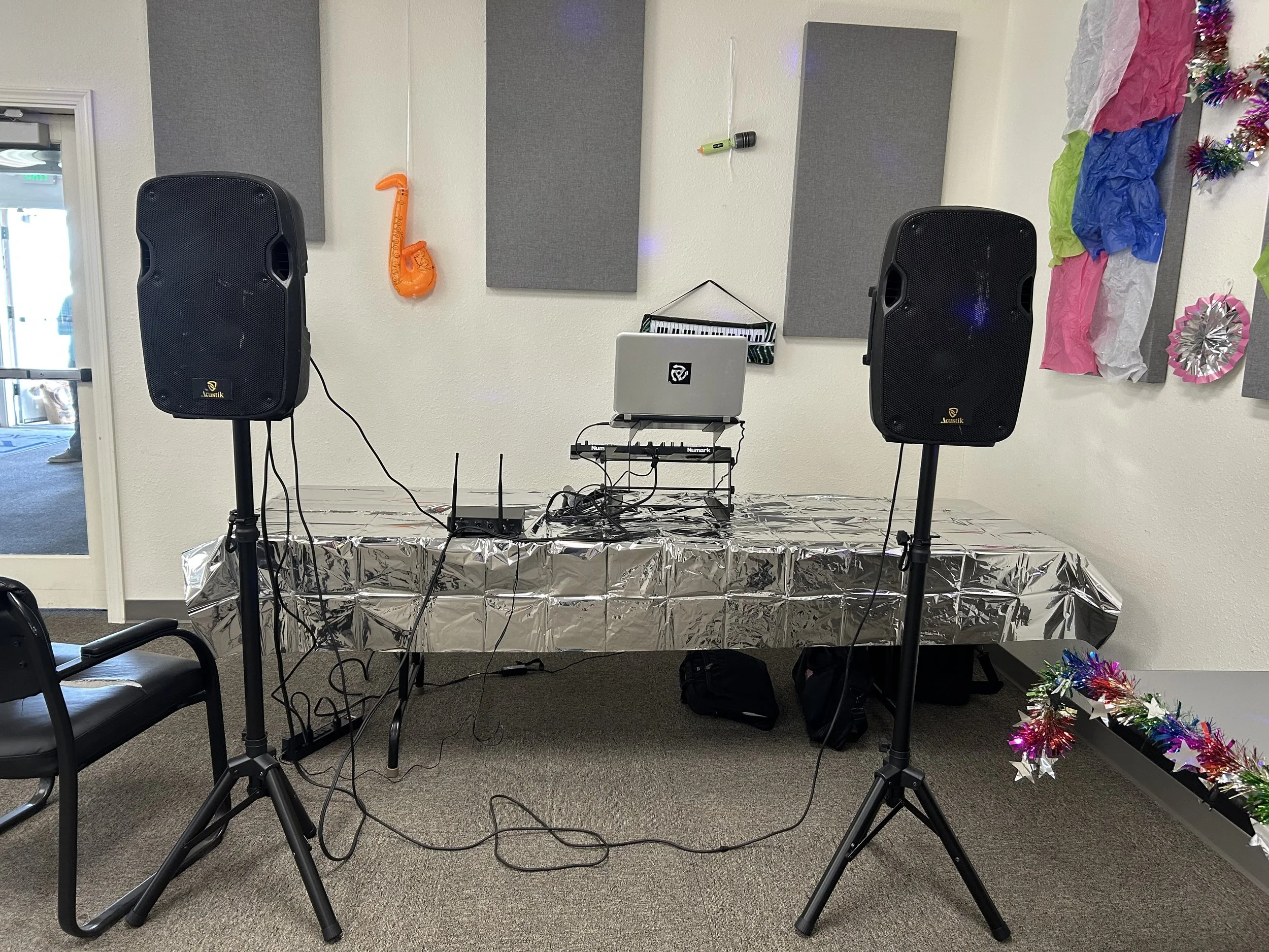 DJ setup with speakers, laptop, and sound equipment on a table, decorated with colorful tinsel and party decorations in a room.