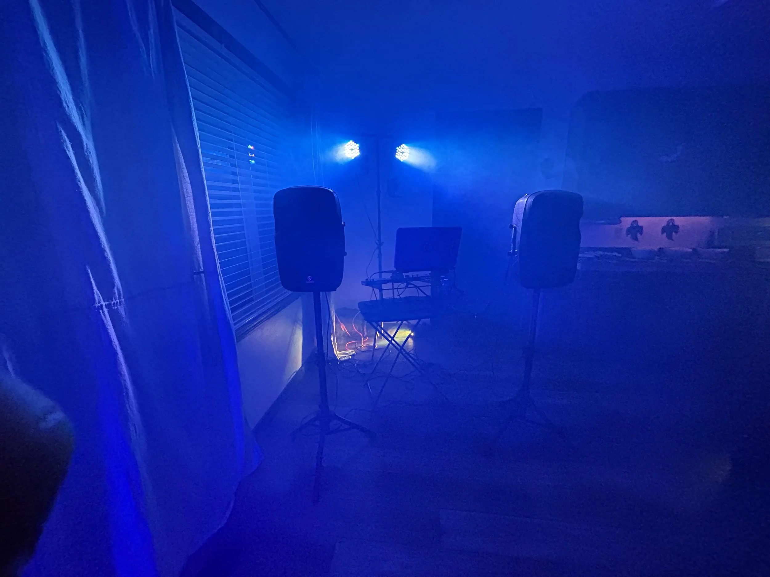 Dimly lit room with blue stage lights, two large speakers on stands, a chair, a small table with audio equipment, and a laptop, suggesting a music or DJ setup.