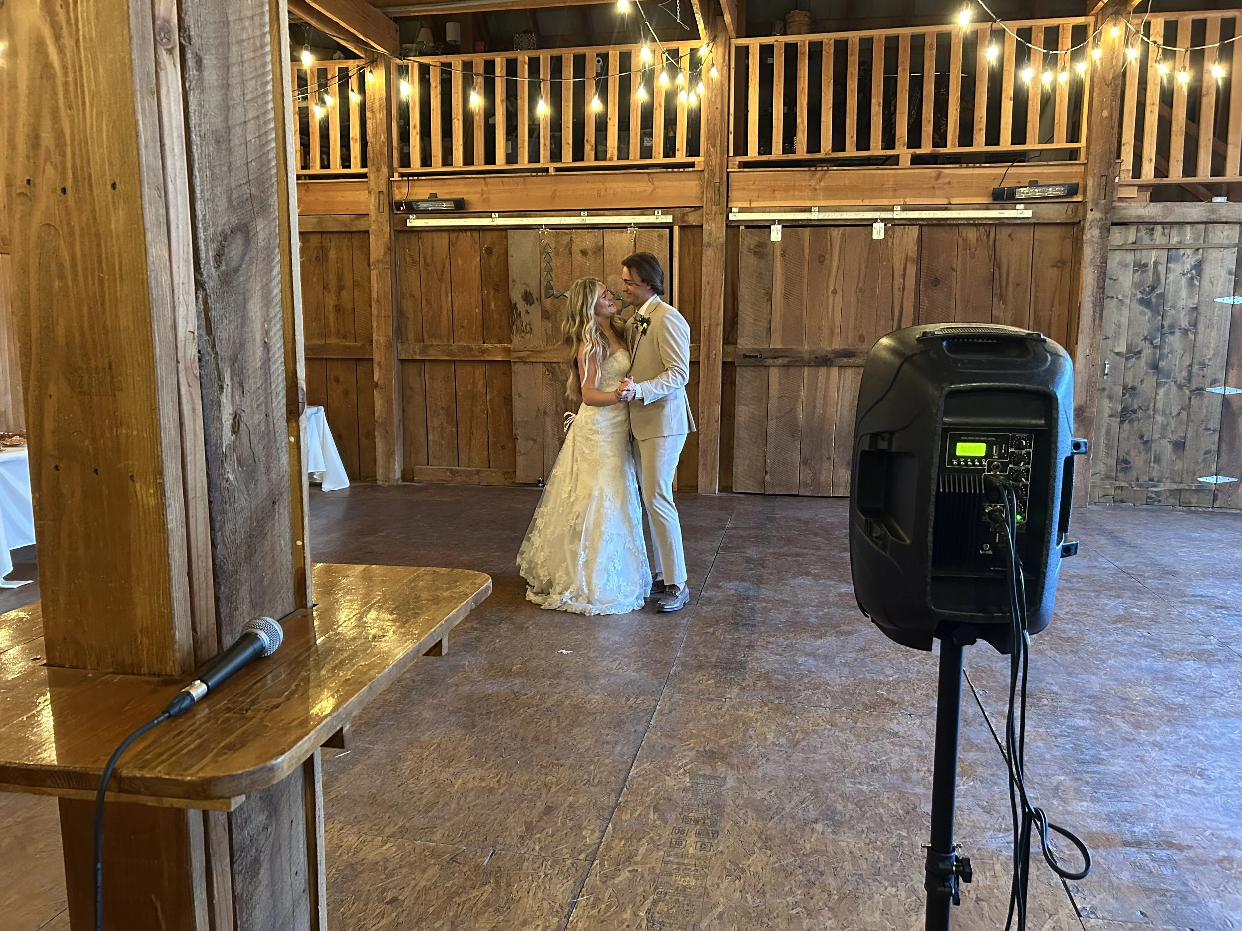 A bride and groom dancing in a rustic wooden barn with string lights overhead.