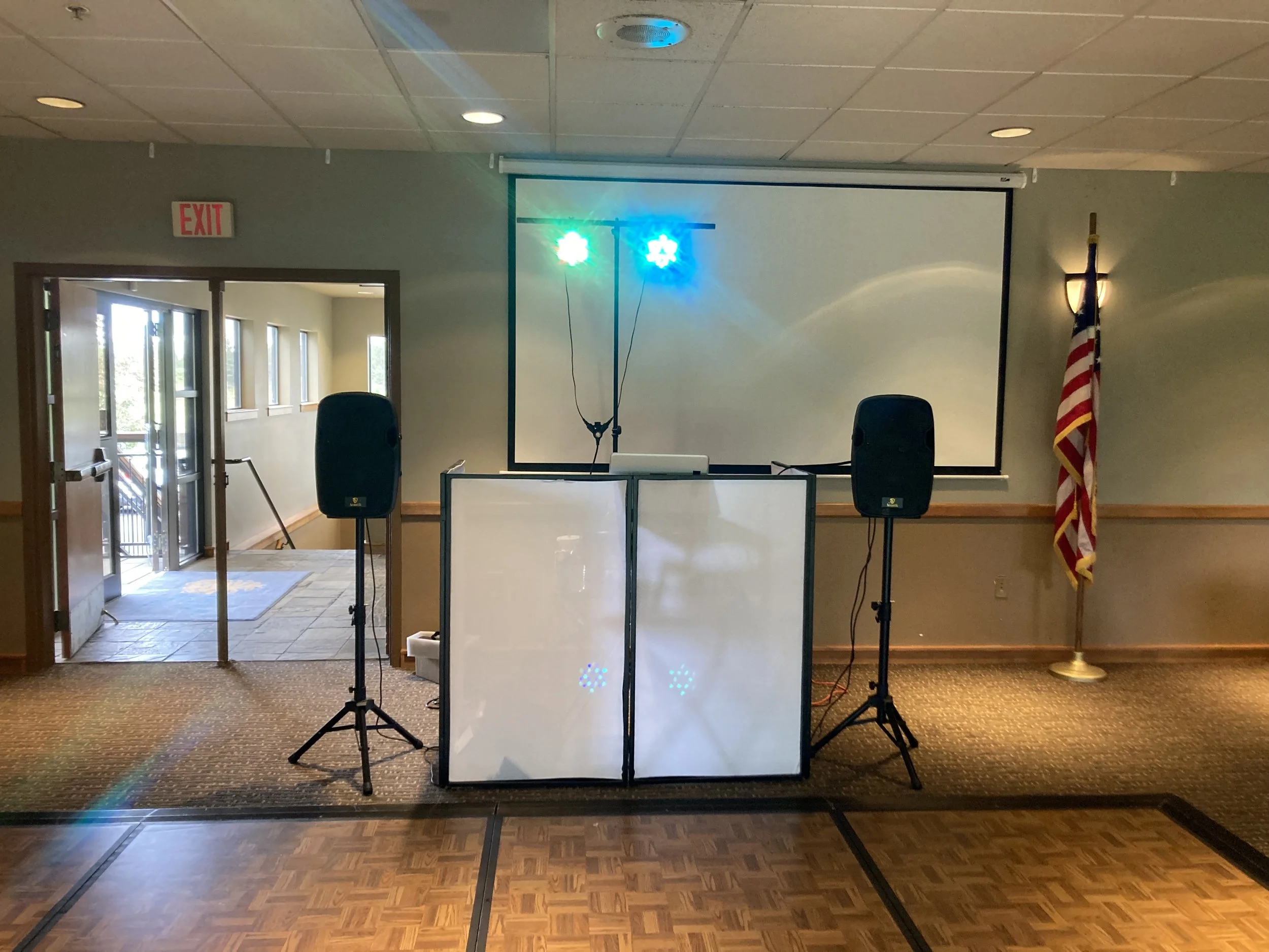 Event setup with two speakers on stands, a DJ booth with lighting, a projection screen behind, an American flag on a pole to the right, and an exit door to the left.