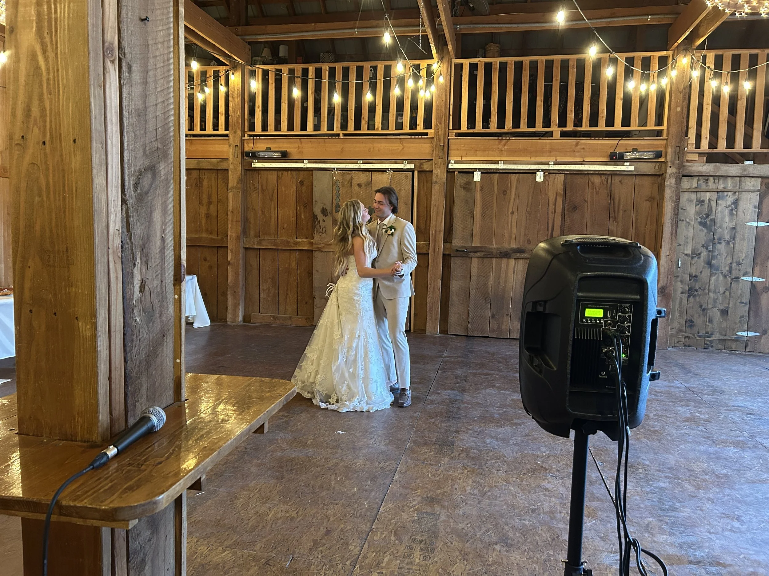 A bride and groom dancing in a rustic barn with wooden walls and string lights overhead.
