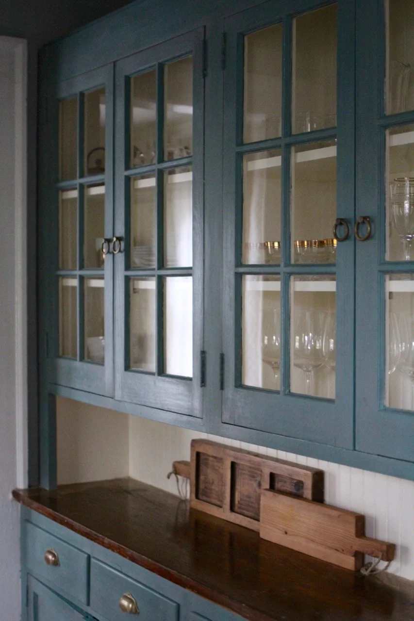 Blue wooden display cabinet with glass doors showcasing glassware inside, placed above a wooden sideboard with brass handles and a wooden tray on top.