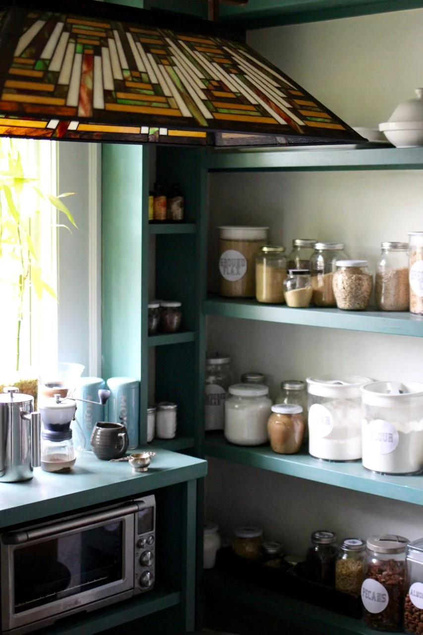 Kitchen shelf filled with various jars of spices, grains, and powders, with a microwave oven below, next to a window with sunlight.