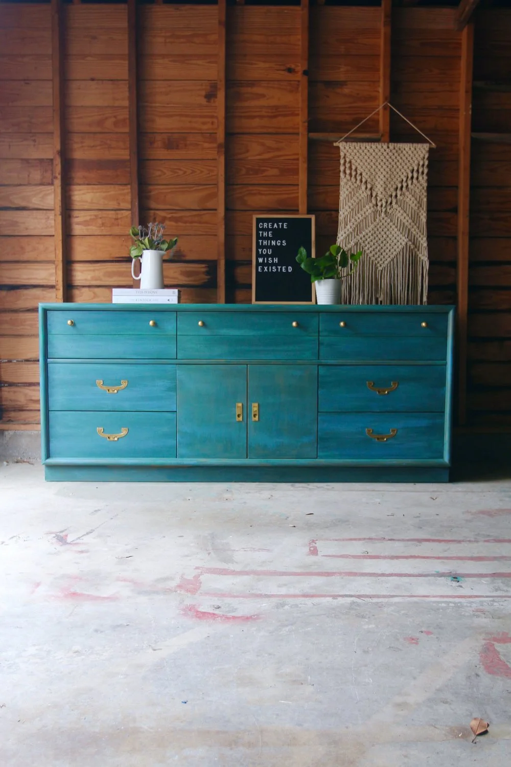 A teal dresser with gold handles against a wooden wall, decorated with a white pitcher of flowers, a book, a black letter board with the quote "Create the things you wish existed," a potted plant, and a hanging macrame wall hanging.
