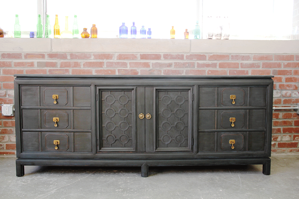 A black sideboard with gold handles, set against a brick wall with glass bottles on a shelf above.