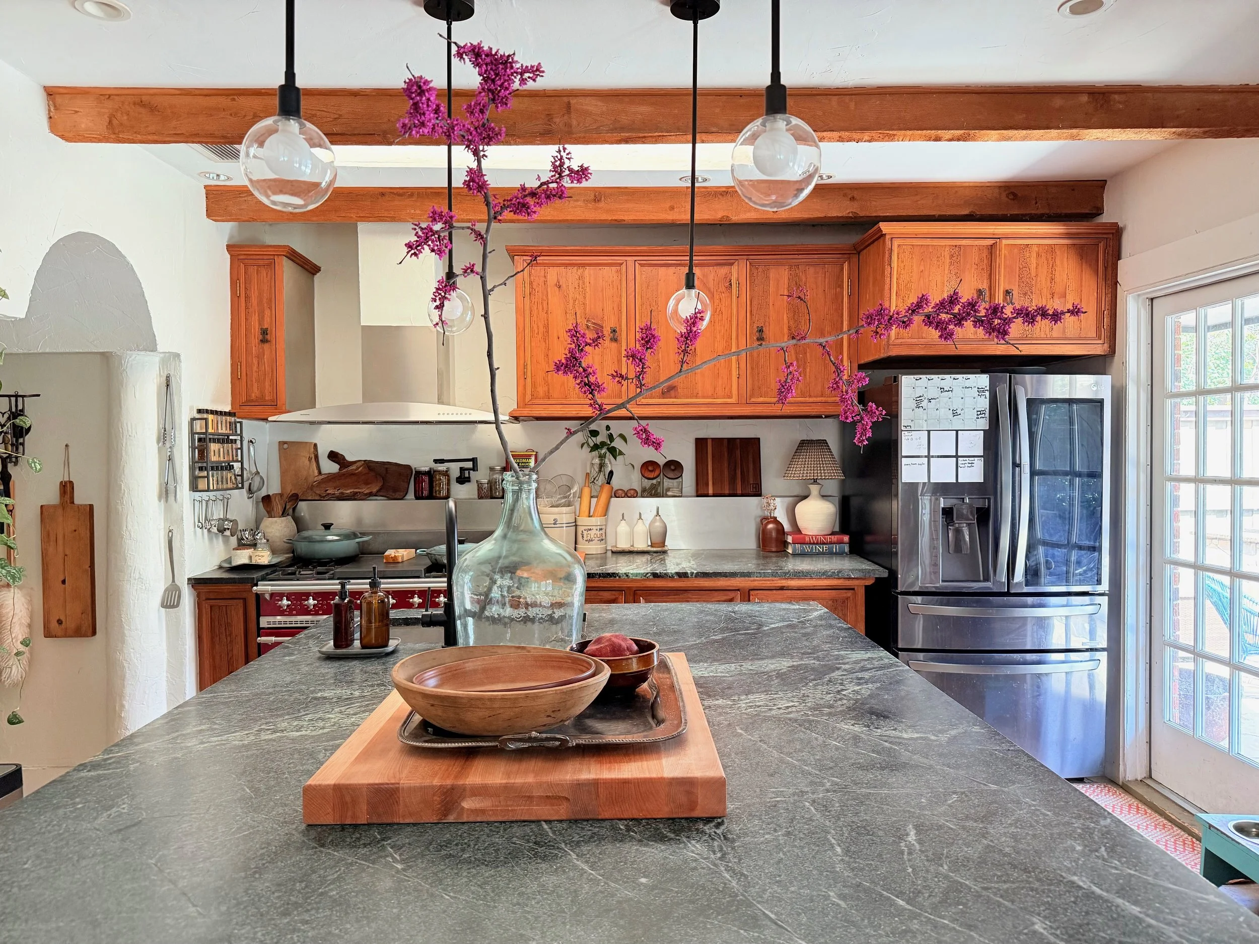 A kitchen with wooden cabinets, a stainless steel refrigerator, and a green marble countertop. There is a decorative branch with purple flowers in a glass bottle hanging from the ceiling. The kitchen has pendant lights, wooden beams on the ceiling, and a sliding glass door.