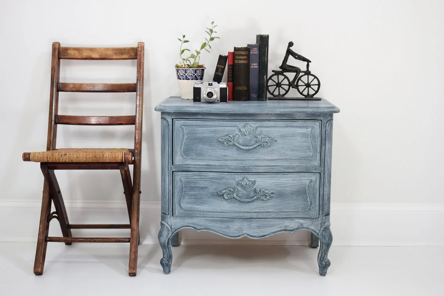 Interior scene with a wooden chair, a blue antique-style nightstand with decorative carvings, a small potted plant, a vintage camera, a stack of books, and a decorative bicycle sculpture.
