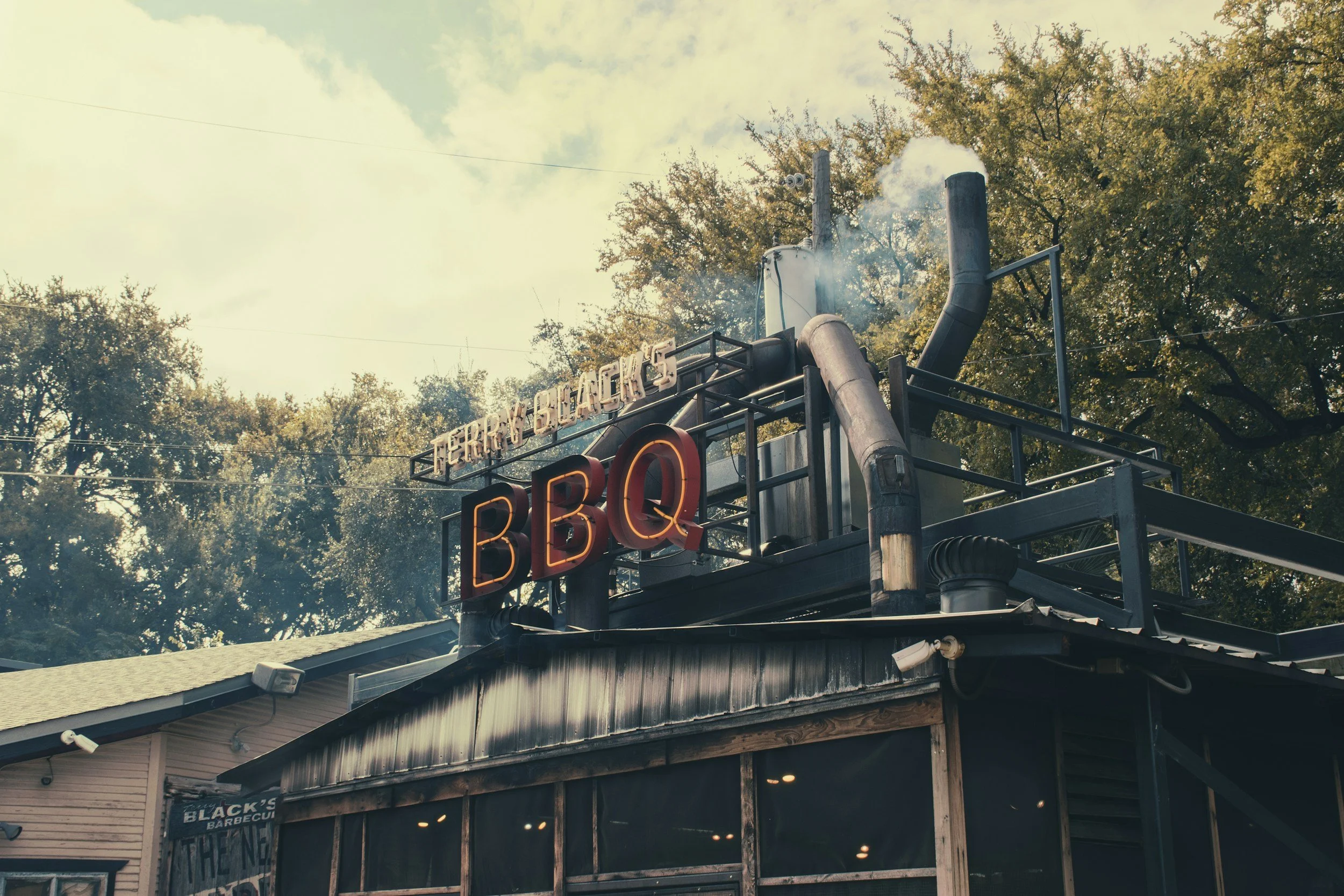 A two-story barbecue restaurant with a neon sign reading 'BBQ' and 'Terry's Beach' on top. There are smoke stacks emitting smoke and a small security camera on the lower roof. The building has wood and metal exterior siding, and there are trees in the background with a partly cloudy sky.