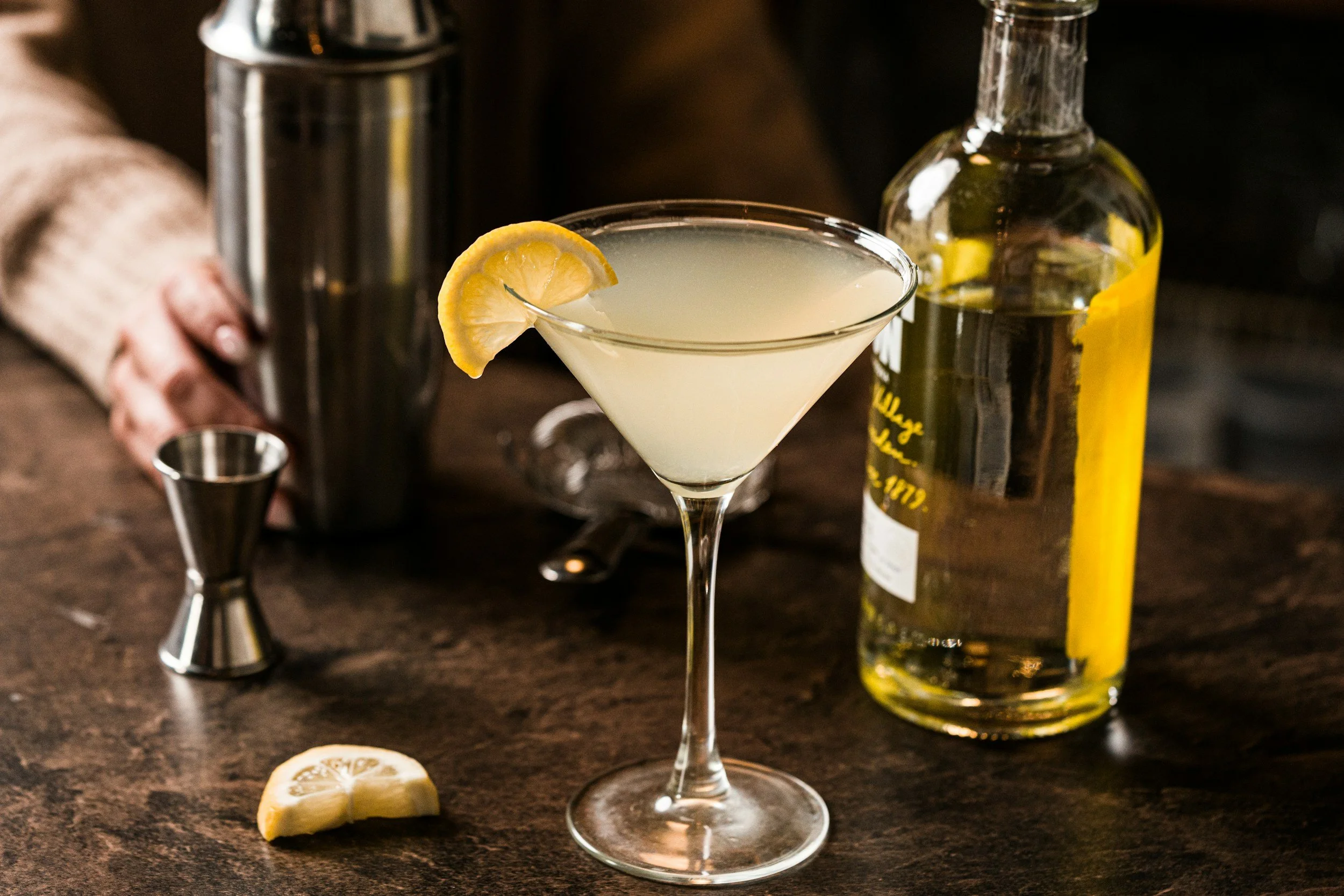 A lemon drop cocktail garnished with a lemon wedge on a dark wooden bar counter, surrounded by cocktail tools and a bottle of lemon-flavored liquor.