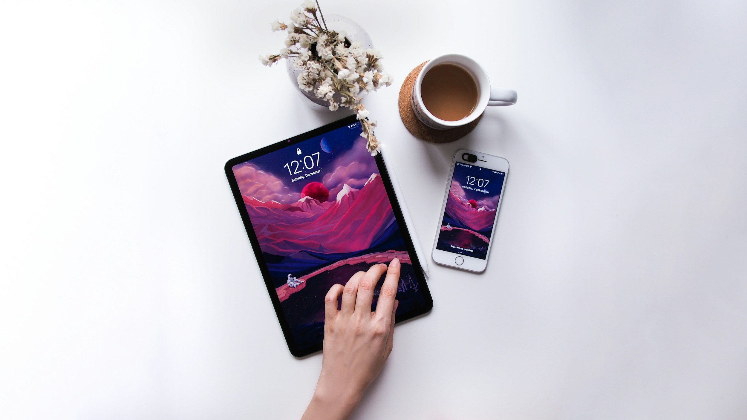 Schermo di un tablet e di uno smartphone con il tema di montagne e cielo sereno, una mano che tocca il tablet, una tazza di caffè e un vaso di fiori secchi sul tavolo bianco.