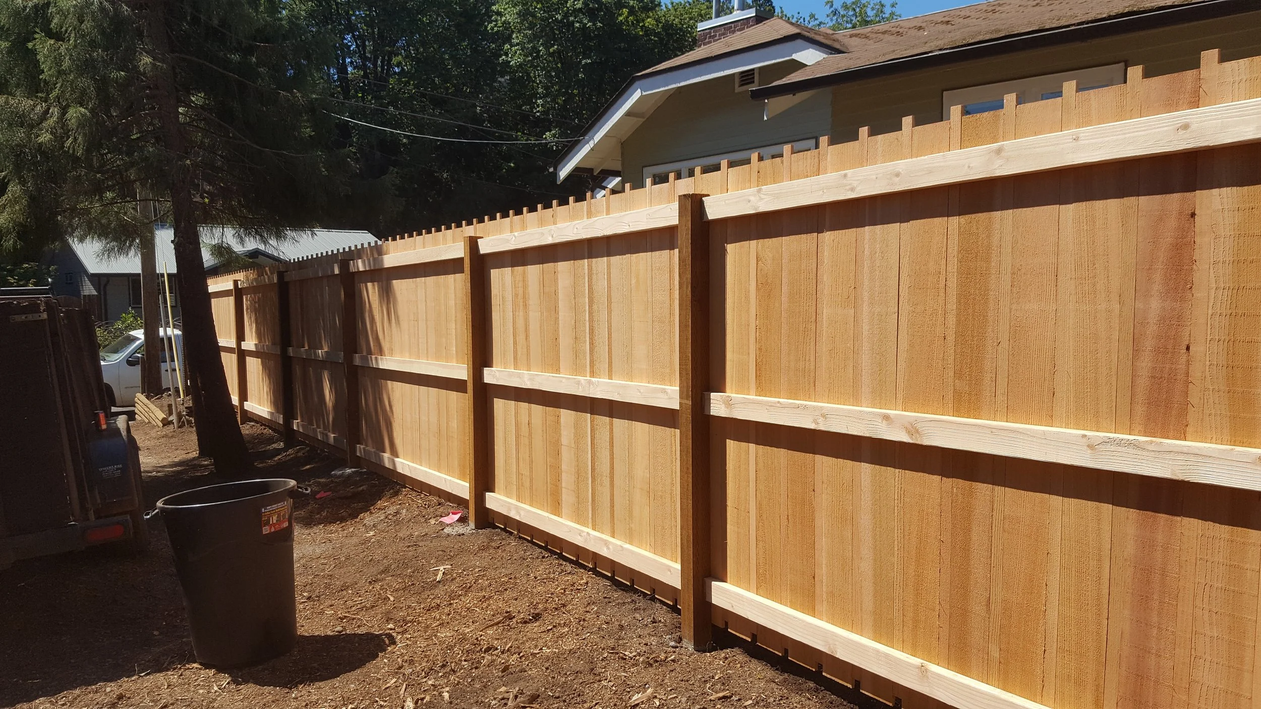New wooden privacy fence installed along a backyard with a house in the background. The fence is tall, made of vertical planks with horizontal support beams, and running parallel to the house.
