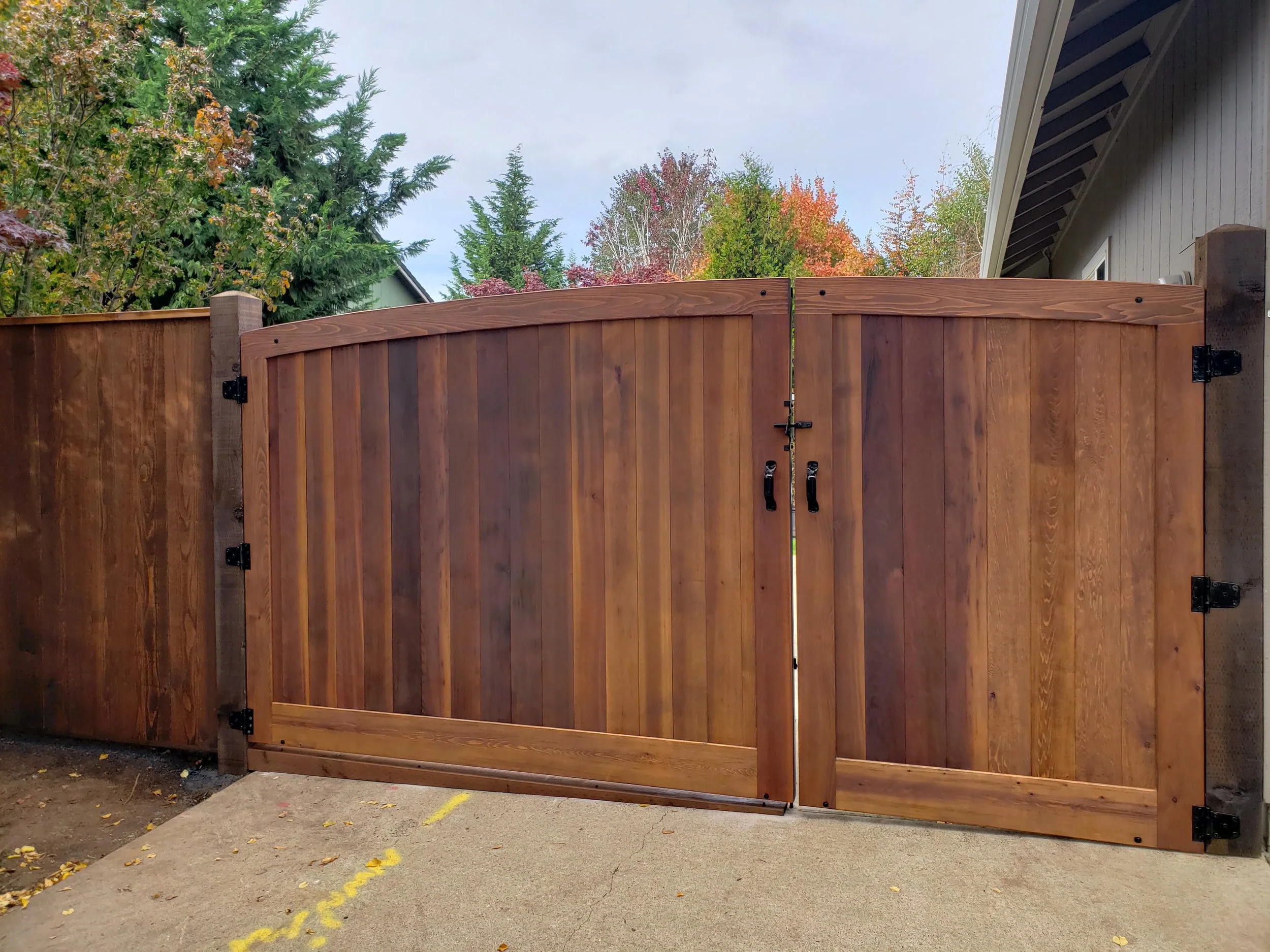 A wooden double gate with black hardware, part of a wooden fence, in front of a house with gray siding. There are trees with autumn foliage in the background and a concrete driveway in the foreground.