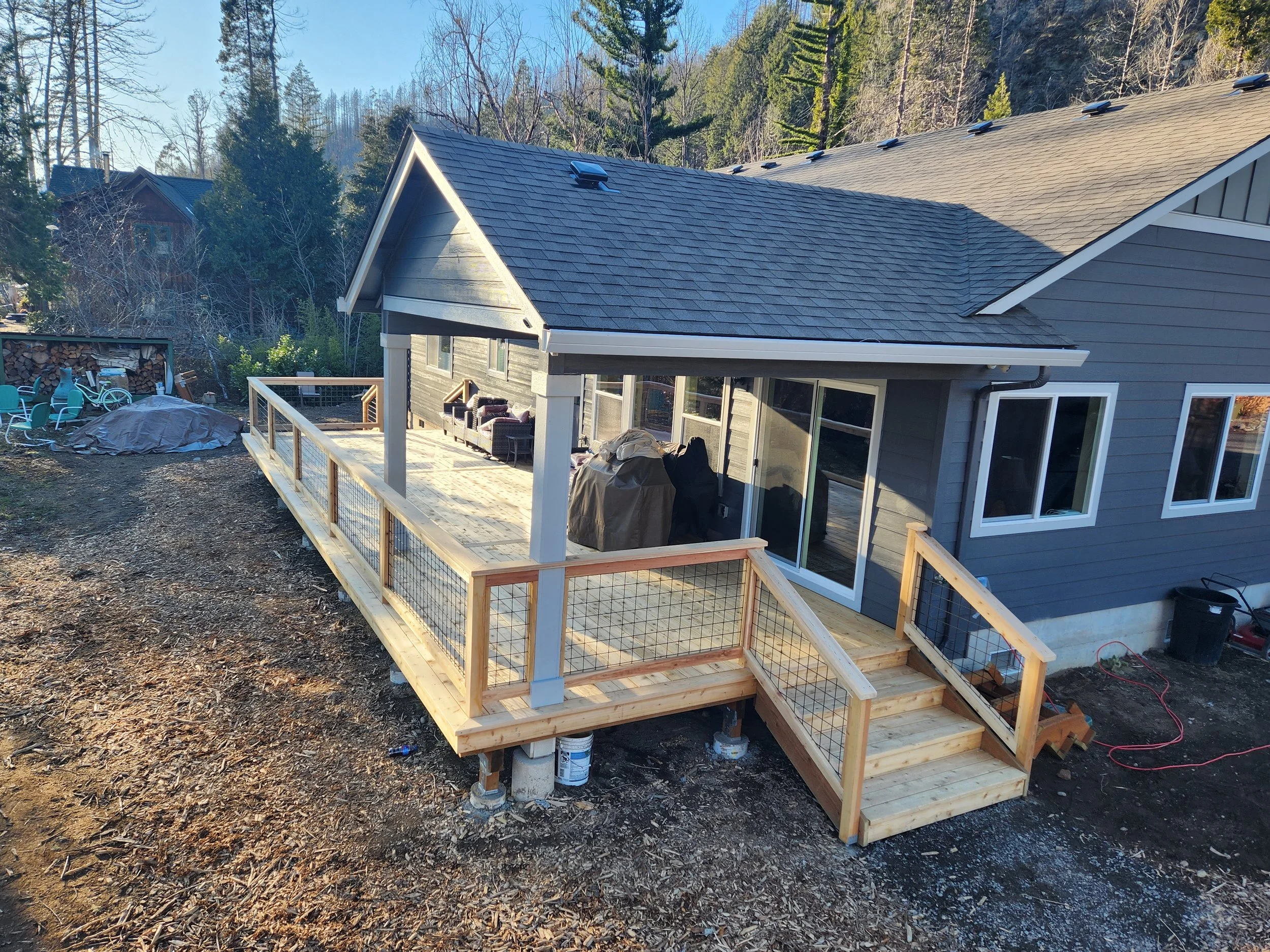 Newly built wooden deck with railing attached to a gray house with sliding glass doors, in a backyard surrounded by trees, dirt ground, outdoor furniture covered with tarp, and construction materials.
