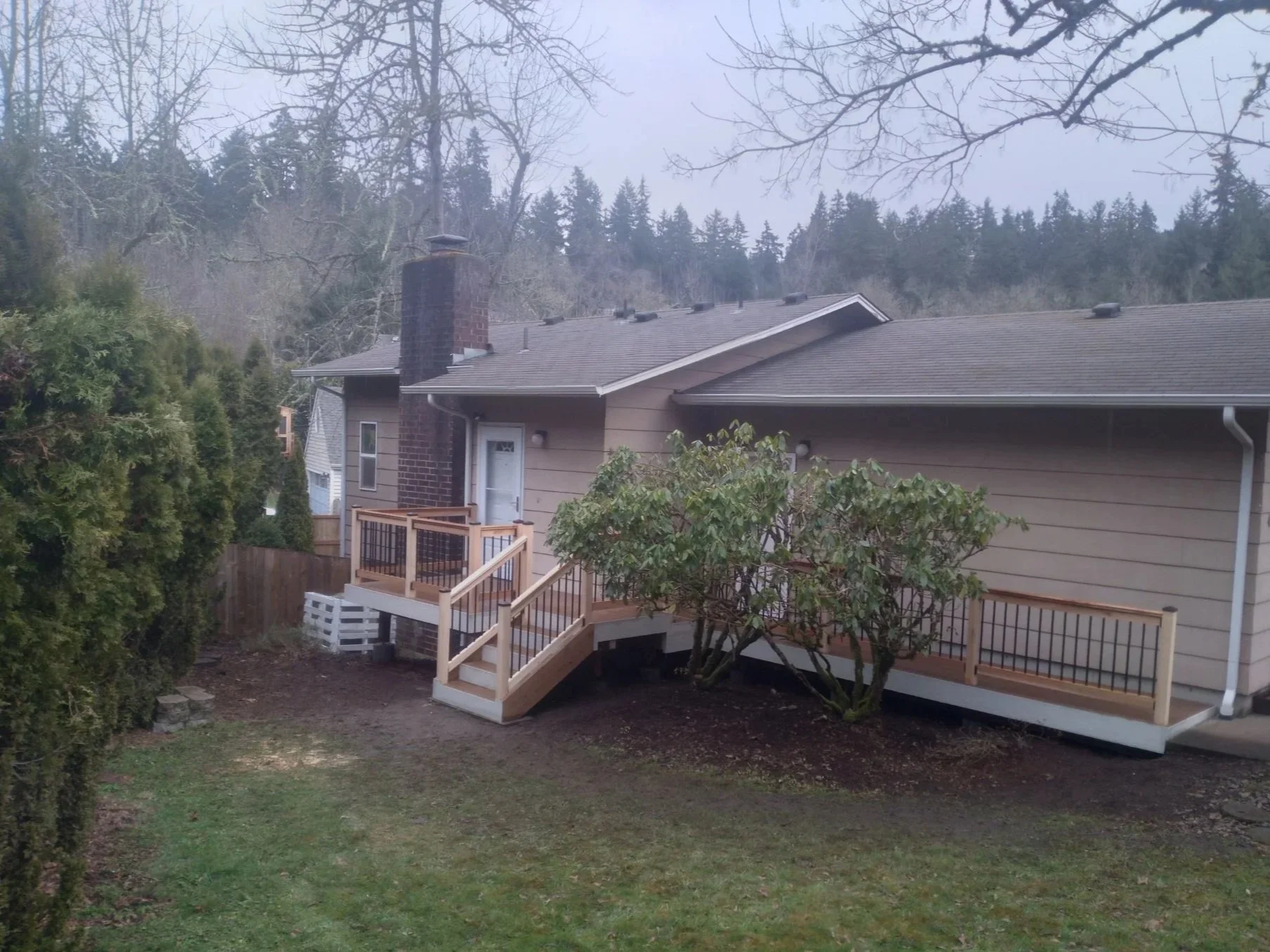 Backyard view of a house with a wooden deck, stairs, and railings, a bush and trees surrounding the yard, and forested hills in the background.