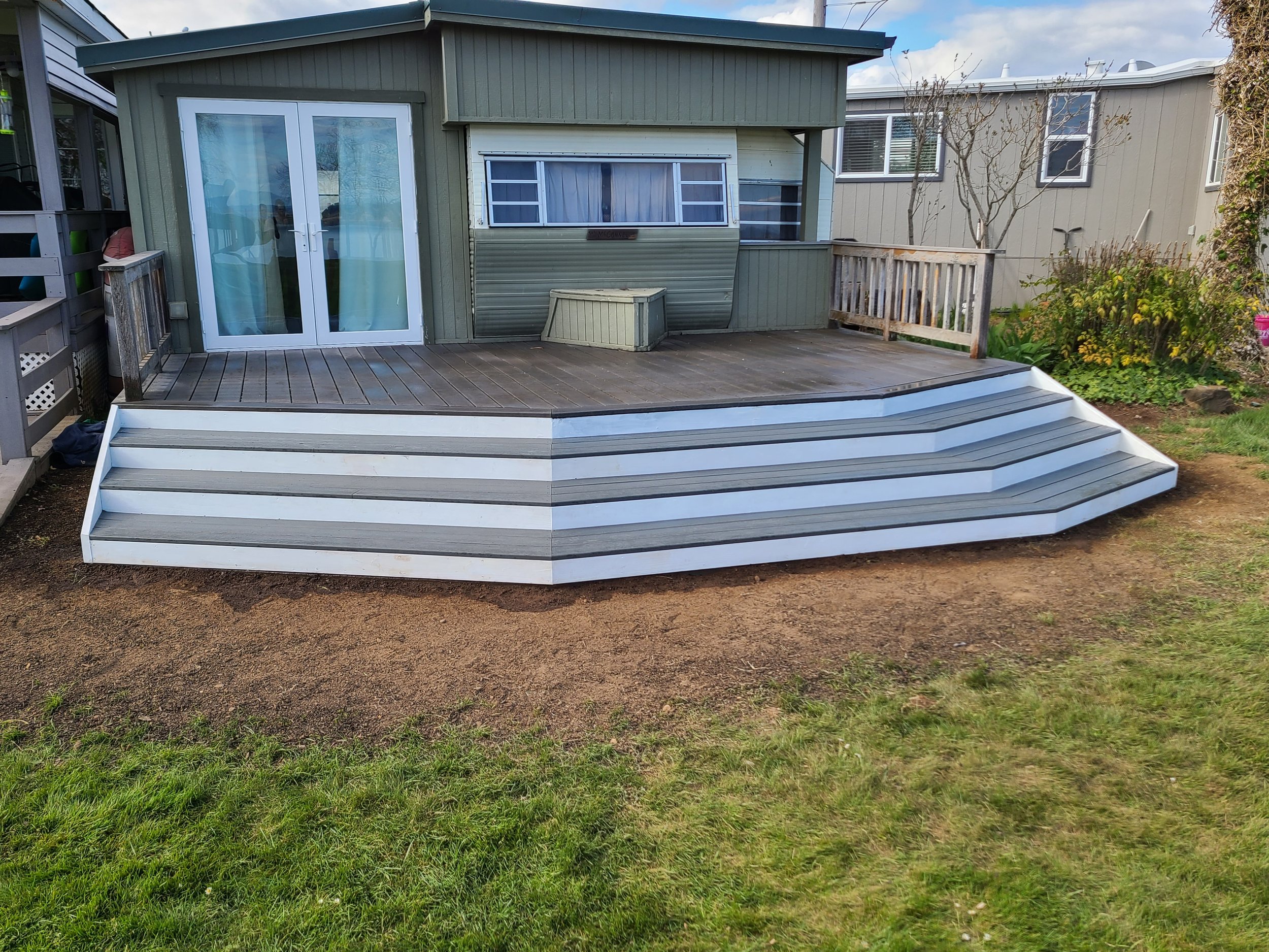Exterior view of a backyard showing a wooden deck with steps leading down to dirt and grass, attached to a green house with sliding glass doors and a window, and neighboring houses in the background.