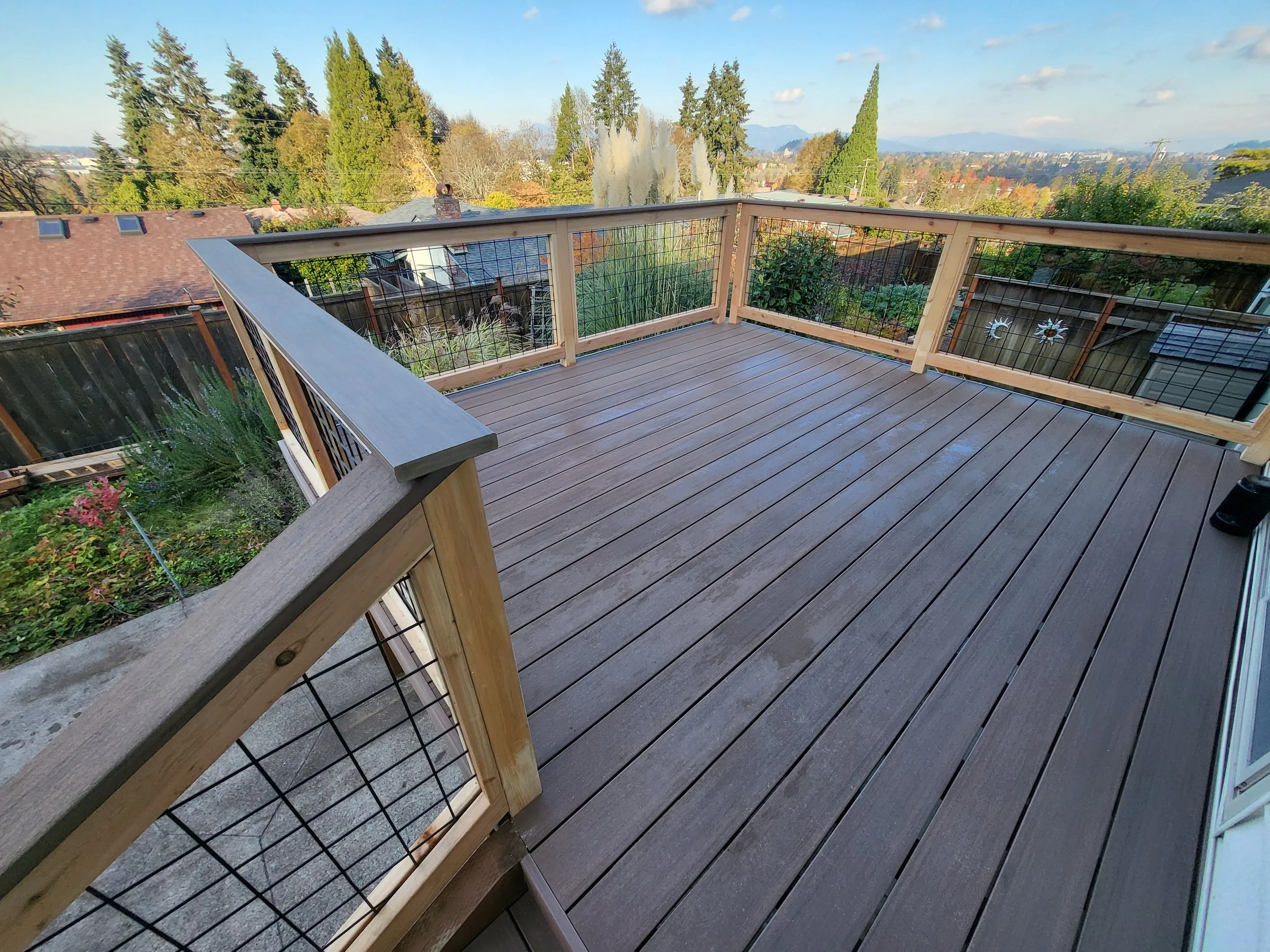 View of a wooden deck with a railing overlooking a suburban neighborhood with trees, houses, and distant mountains in the background.