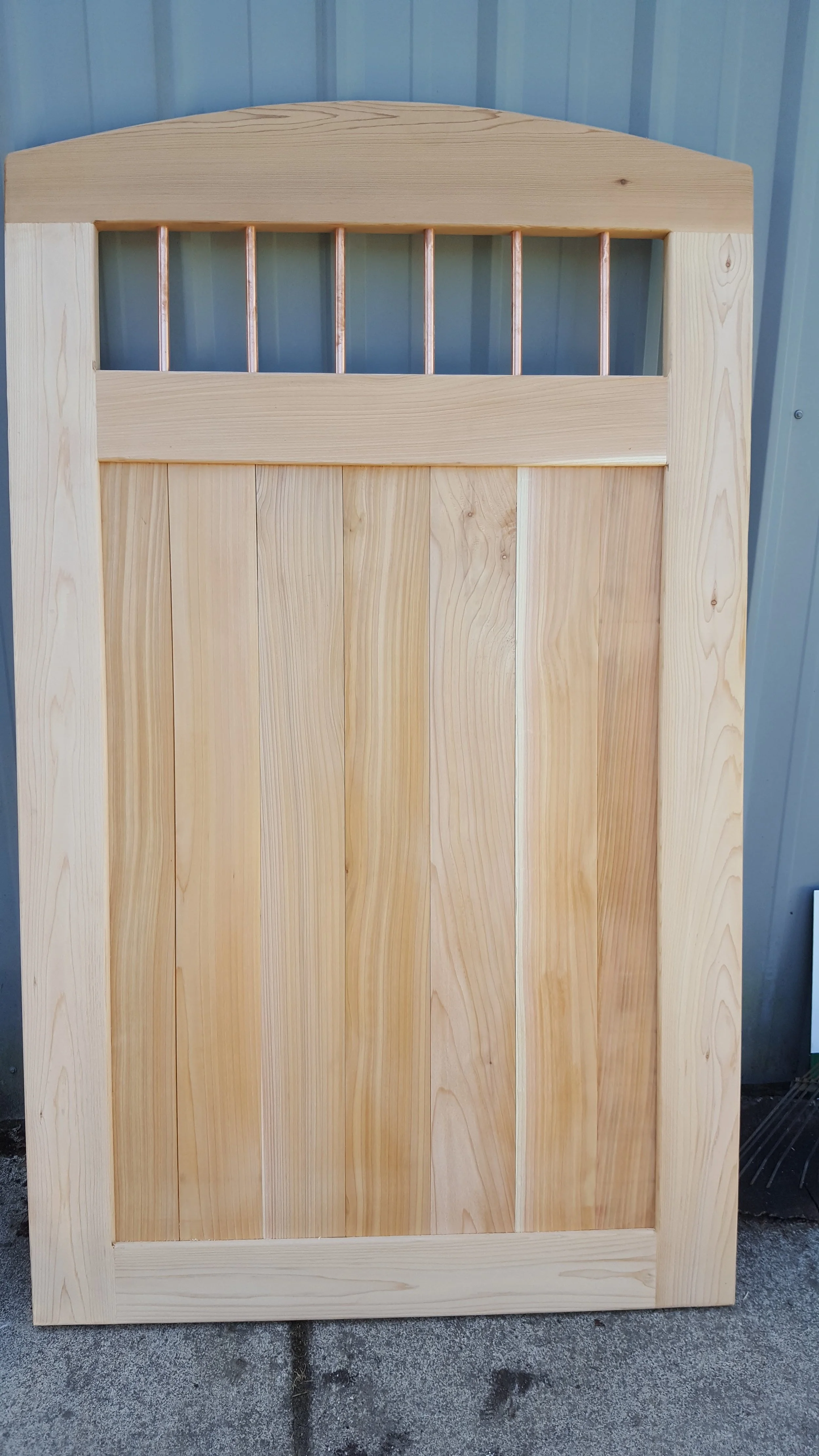 A wooden door with vertical panels, topped with a horizontal bar and a small window with three vertical bars, set against a corrugated metal wall.