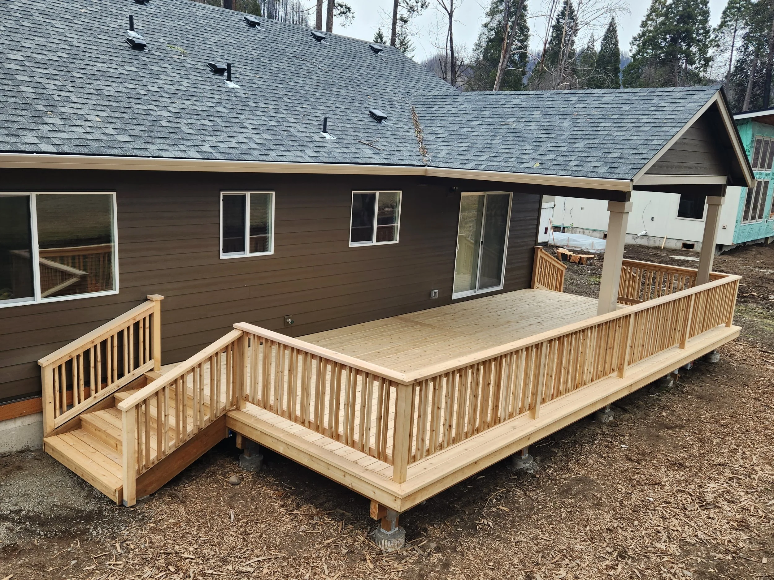 New wooden deck with railing attached to the back of a house with a brown exterior and a gray shingled roof, in a wooded area.