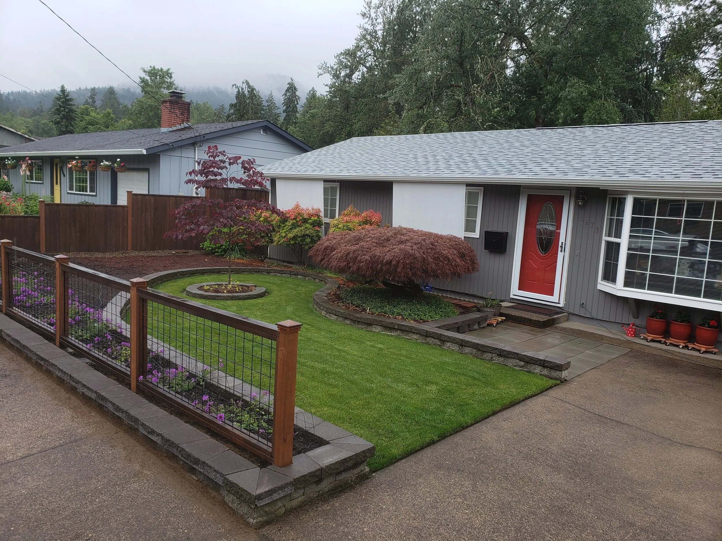 Front view of a gray house with a red door, manicured lawn, and landscaped garden with small trees and flowers, surrounded by a wooden and metal fence on a rainy day.