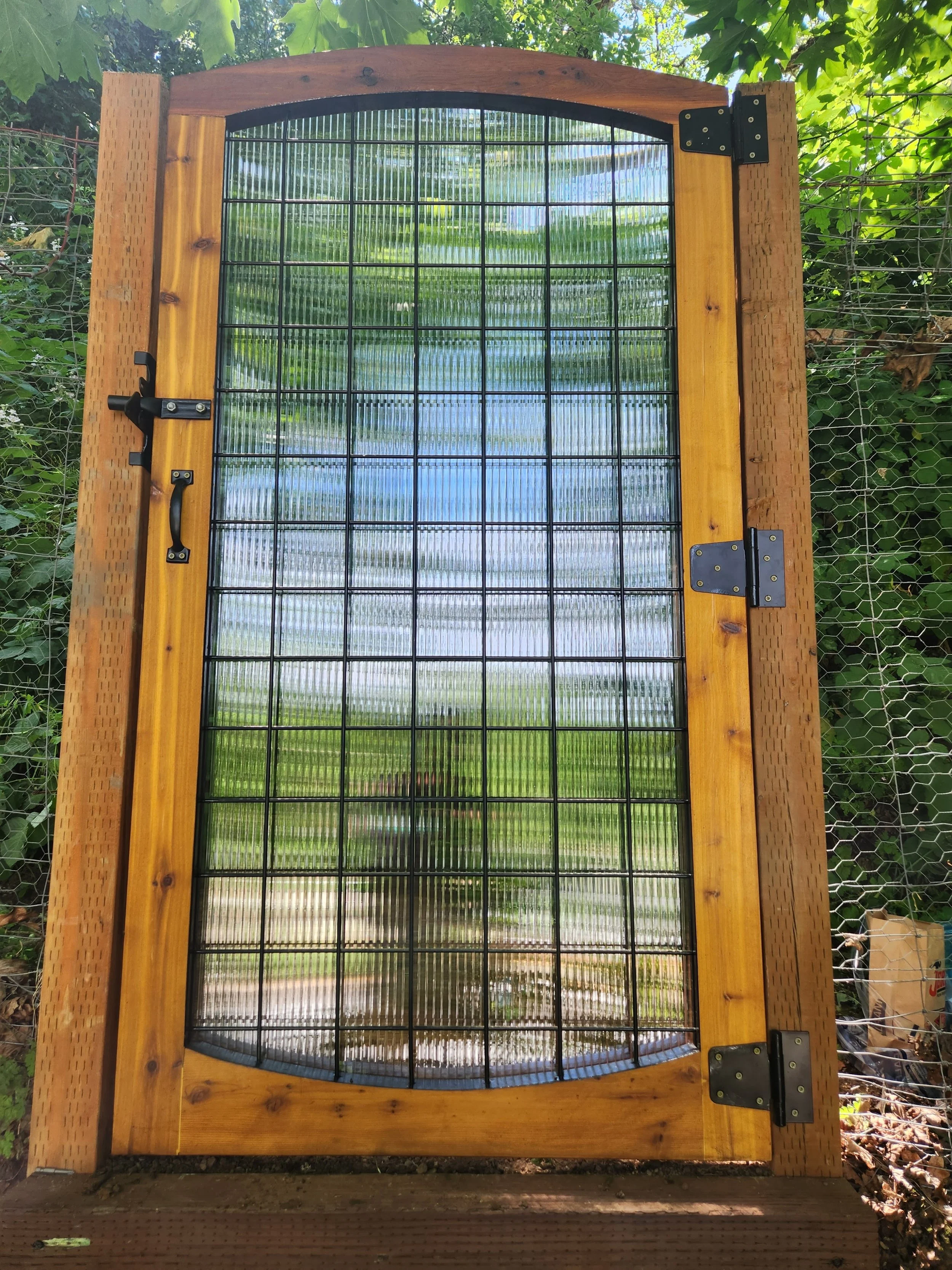 A wooden and metal gate with a grid design, installed outdoors against a green leafy background.