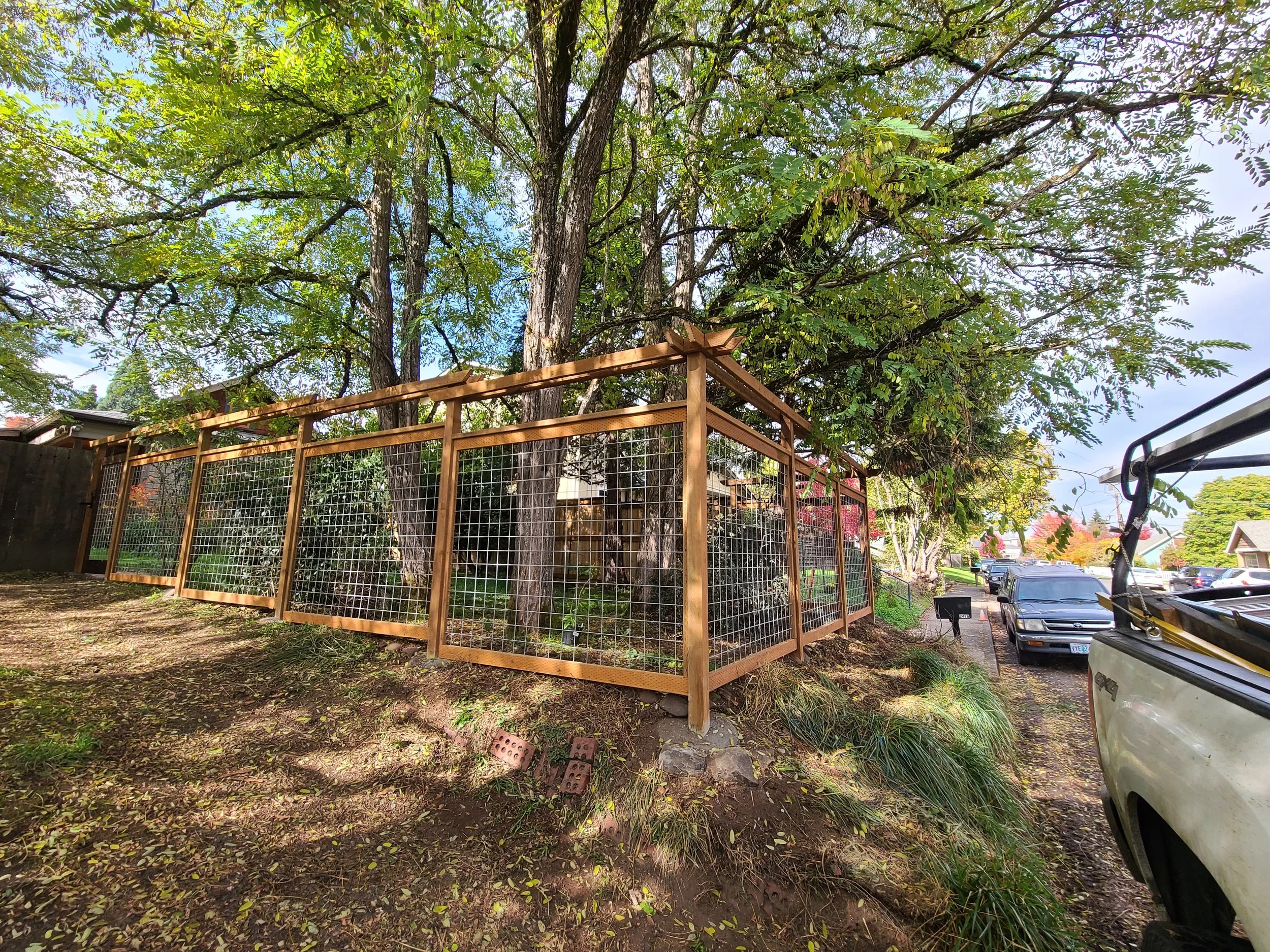 Wooden fenced enclosure in a yard with trees, plants, and parked cars nearby.