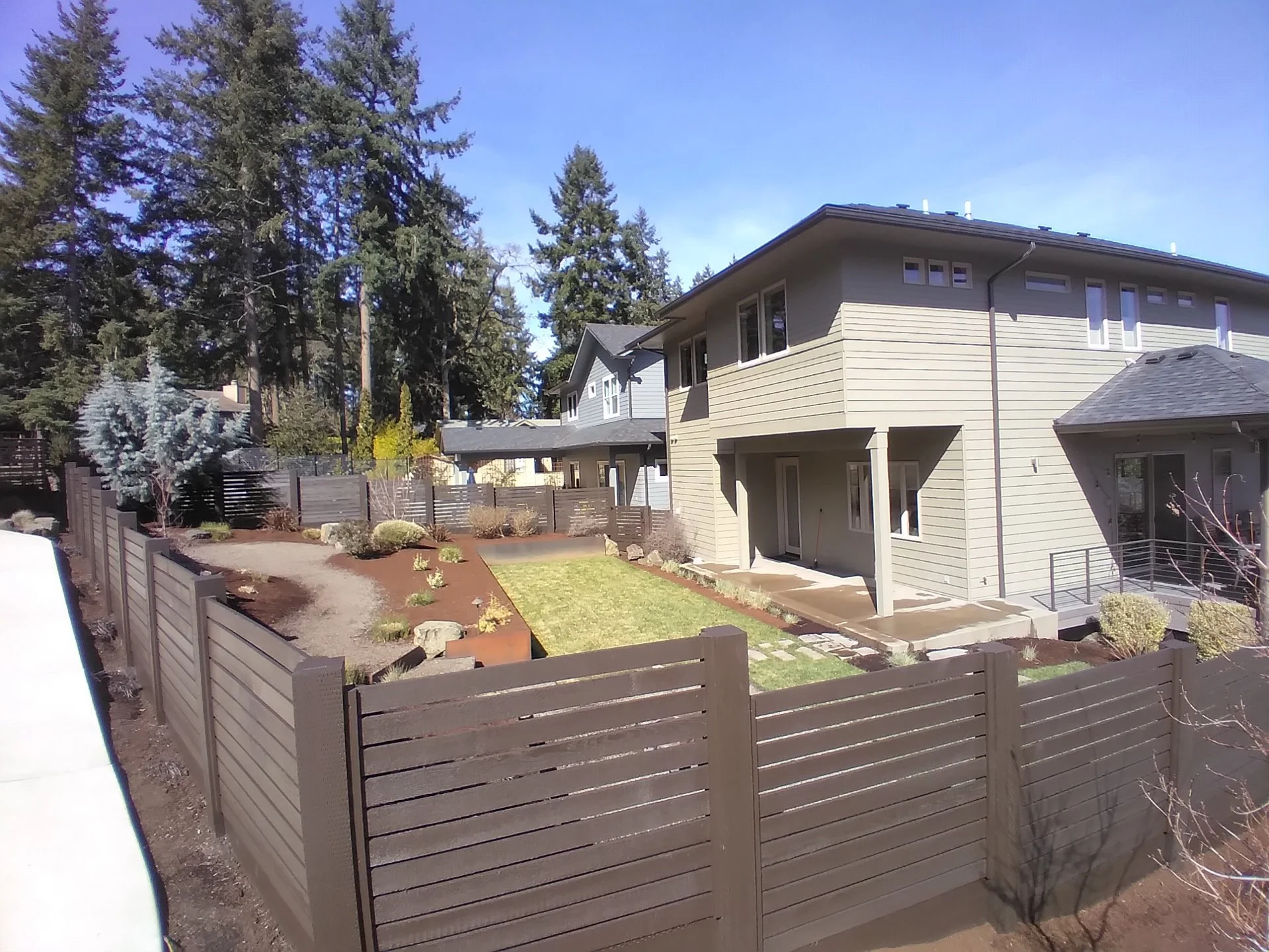 Rear view of a modern house with a fenced backyard, small lawn, and landscaped garden with trees and shrubs during daytime.