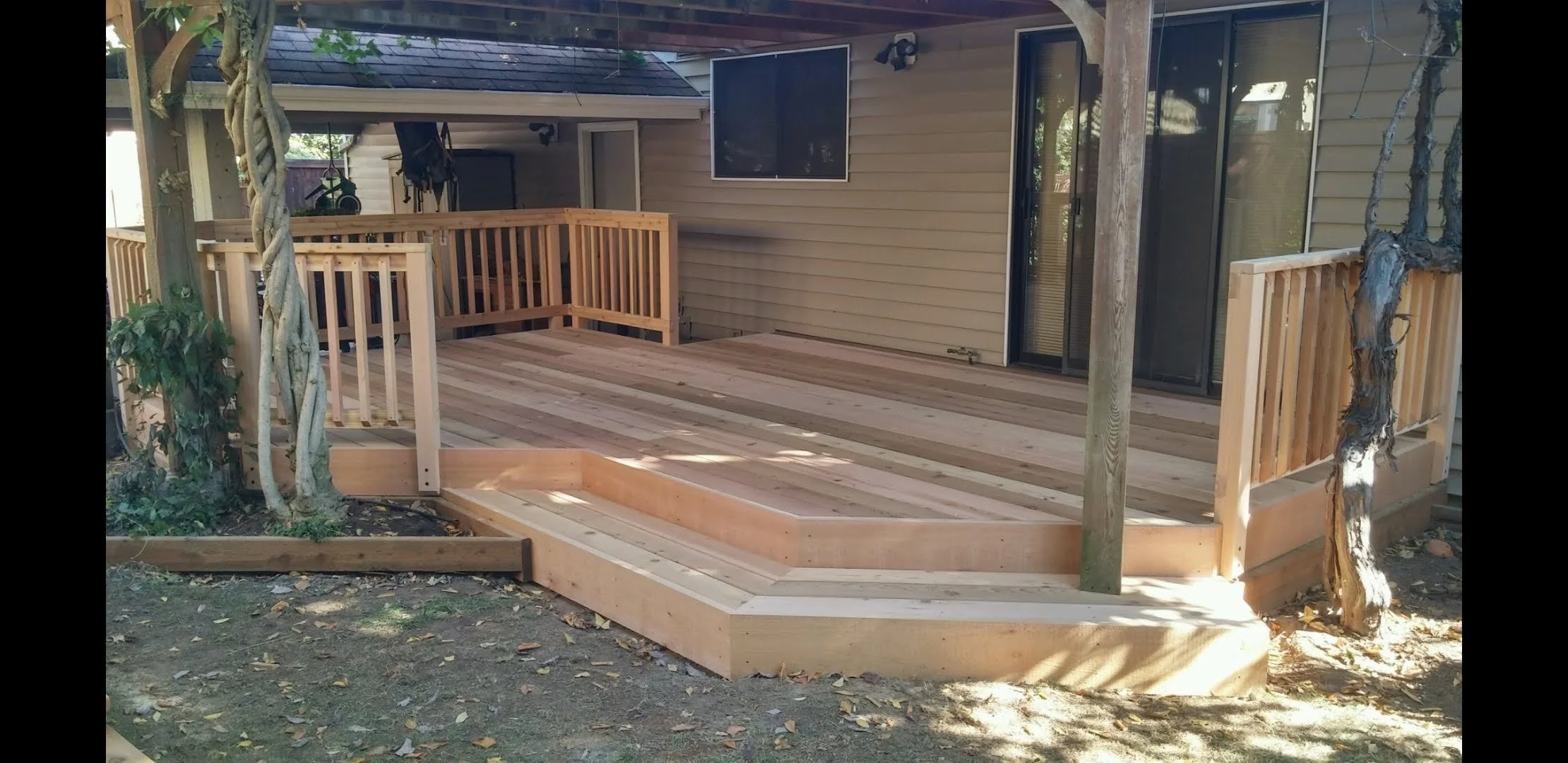 Freshly built wooden deck with railing, attached to the back of a house, with a sliding glass door and window, situated in a backyard with trees and fallen leaves.