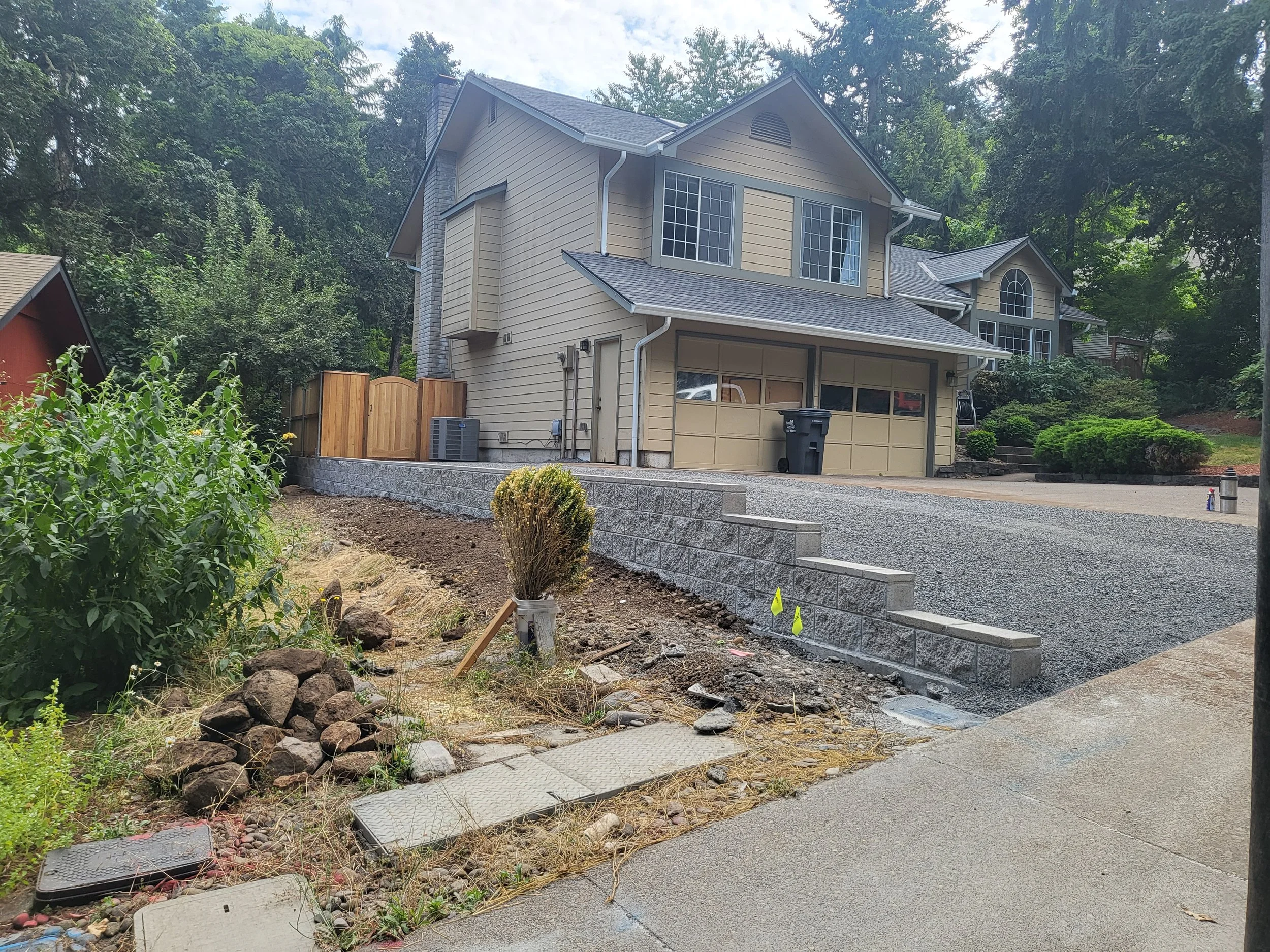 Residential front yard with a new landscaped retaining wall, a gray driveway, and a beige house with multiple windows and a garage, surrounded by trees and bushes.