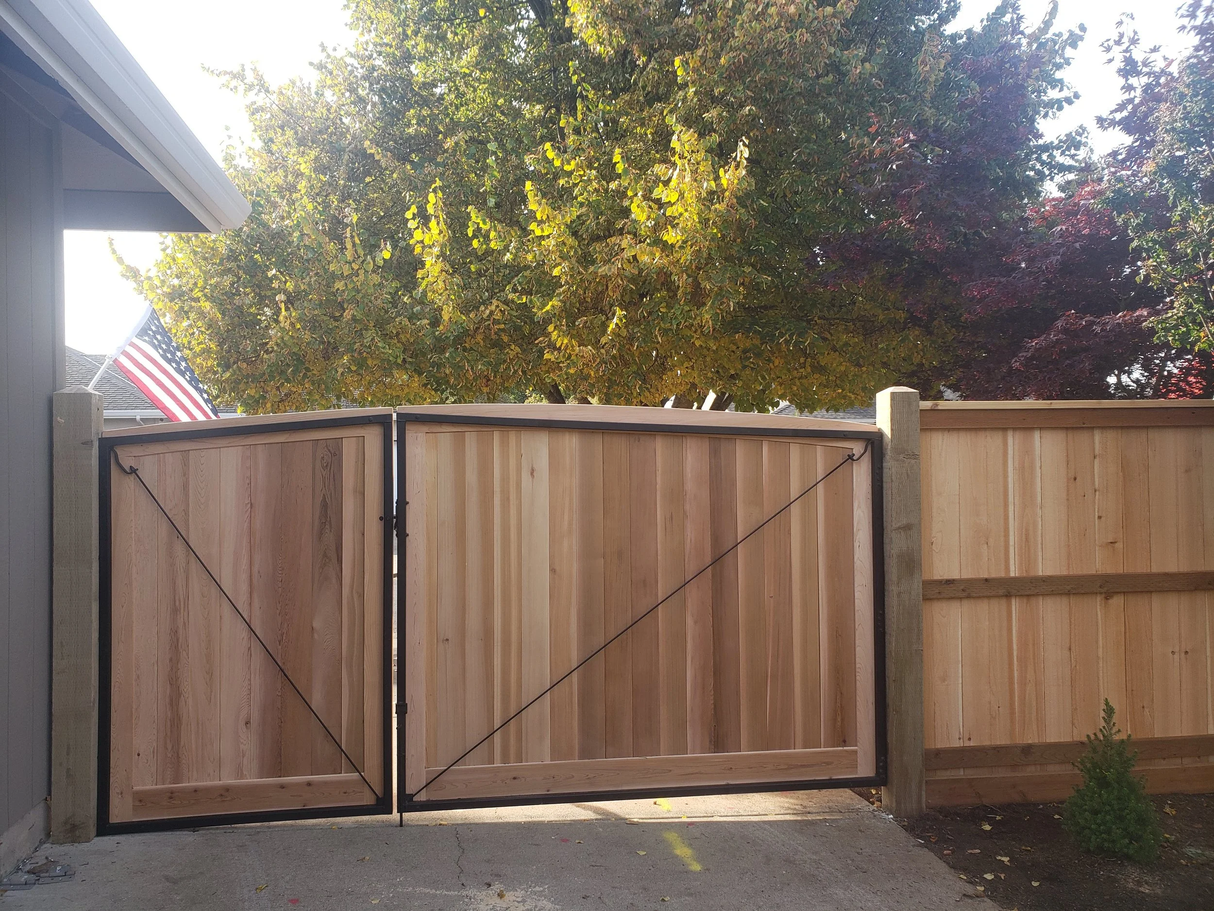 A wooden gate attached to a fence on a concrete driveway, with trees and an American flag visible in the background.