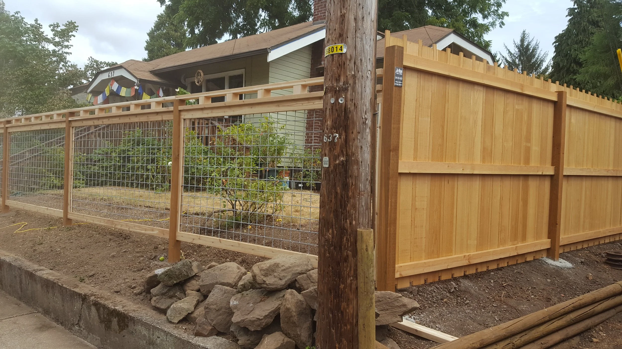 A residential front yard enclosed by a new wooden fence with a wire section, with a house in the background, trees, and construction materials on the ground.