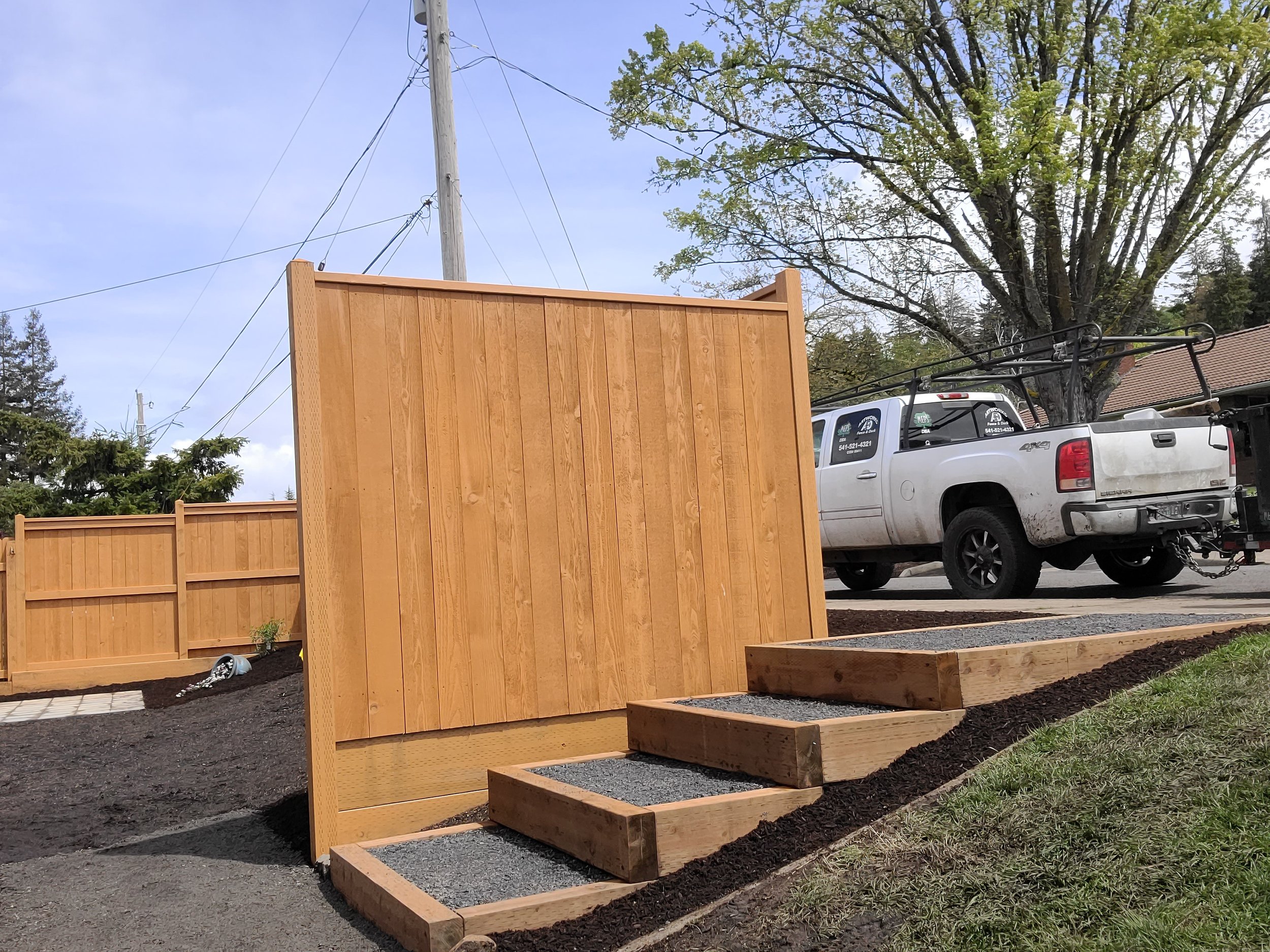 A backyard with a tall wooden privacy fence, a small set of wooden stairs with gravel in the steps, a white pickup truck parked on the street, and a large tree with green leaves.