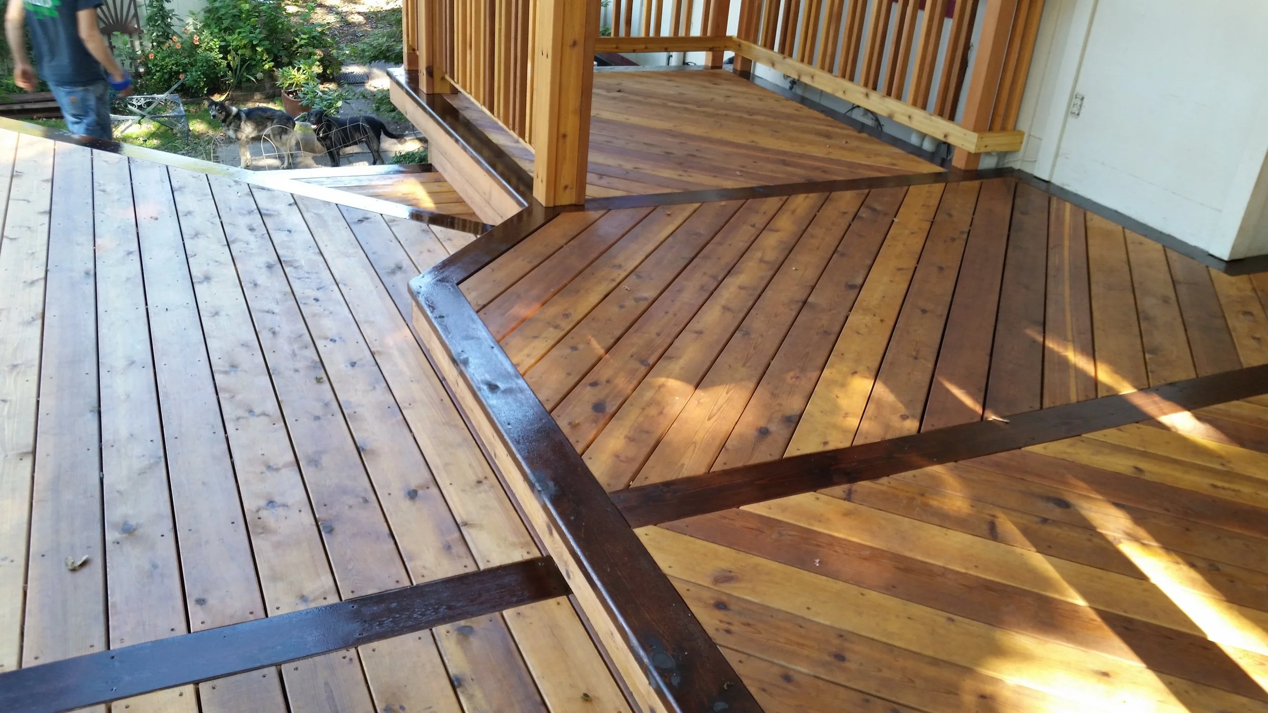 Newly installed wooden deck with areas in different shades of stain, next to a white building with a wooden railing and partially finished structure, with a garden and two cats outside.